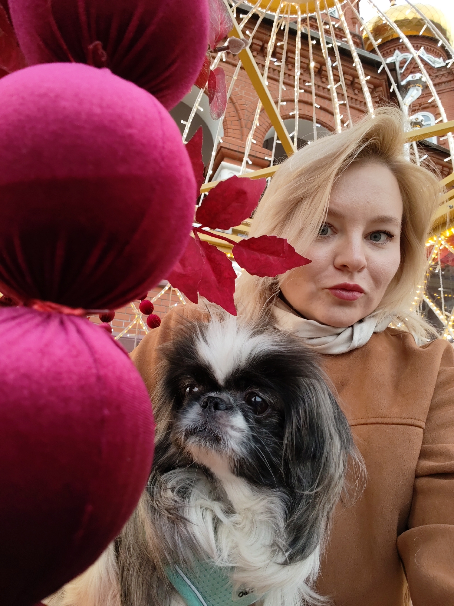 A woman holds a Pekingese dog. The woman is fair-skinned with blonde hair and wears a tan jacket and a cream-colored scarf. The dog has a black and white coat. To the left is a string of large, round, pink decorations. In the background, a red brick building with a gold dome is visible, adorned with a yellow frame and decorative lights. The location is Izhevsk, Russia.