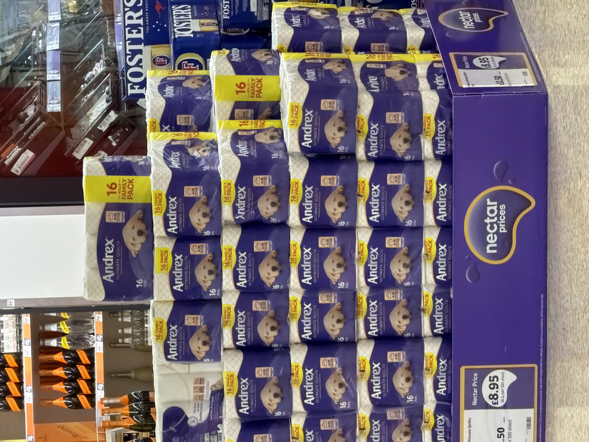 The image shows a stack of Andrex Ultimate Quilts toilet paper, specifically the 16-roll family pack, displayed in a store. The packs are stacked on top of each other, forming a pyramid-like arrangement. A blue "Nectar Prices" promotional box sits beside the toilet paper, indicating a discounted price of £8.95 (compared to the original price of £11.50). The location of the store is suggested by the "Nectar Price" label, which is associated with the Sainsbury's supermarket chain, which may potentially be situated in Pity Me, United Kingdom. The packs of toilet paper feature the Andrex brand name in bold letters and an image of a Labrador puppy. A shelf displaying bottles of alcoholic beverages like Prosecco is located to the left of the toilet paper display. Another brand of lager, Fosters, can be seen in the upper left part of the image.