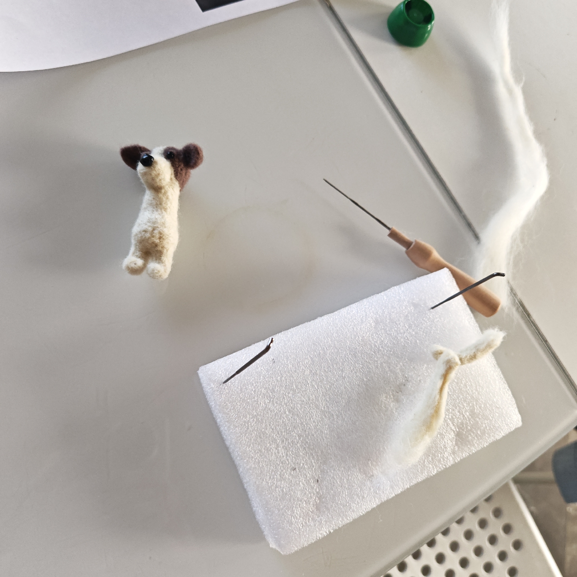 A tabletop scene presents the craft of needle felting. A small, completed felted dog figure stands to the left. A white foam block rests at the lower center, with tufts of wool and two needles embedded within. A felting needle with a light brown handle lies nearby. A length of white wool stretches toward the upper right. A green-capped container is visible in the upper right corner. A partially visible piece of paper is in the upper left. The surface appears to be a light gray or white tabletop. The location is not specified.