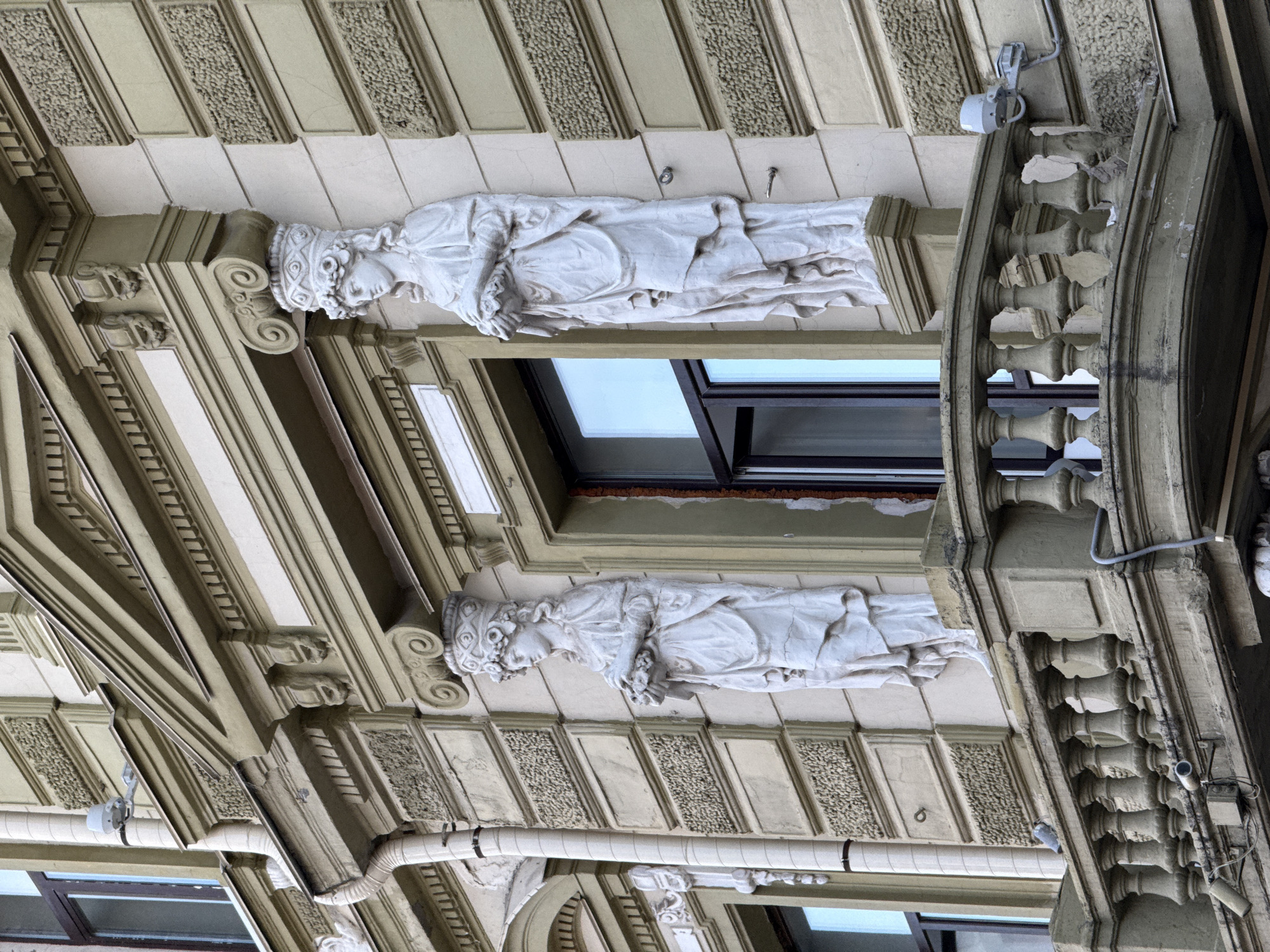 The image shows a section of the facade of a building in Nizhny Novgorod, Russia. Two white stucco sculptures of women in classical attire are positioned on either side of a window. Both sculptures are adorned with elaborate headdresses and are holding bouquets of flowers. They stand atop decorative pedestals. In front of the sculptures, a balustrade of a balcony is visible, with a pigeon perched on its railing. The building's facade is painted in light colors and features decorative architectural elements such as cornices and textured panels. The window to the right of the sculptures is partially open. The overall scene depicts a detail of historical architecture in an urban setting.