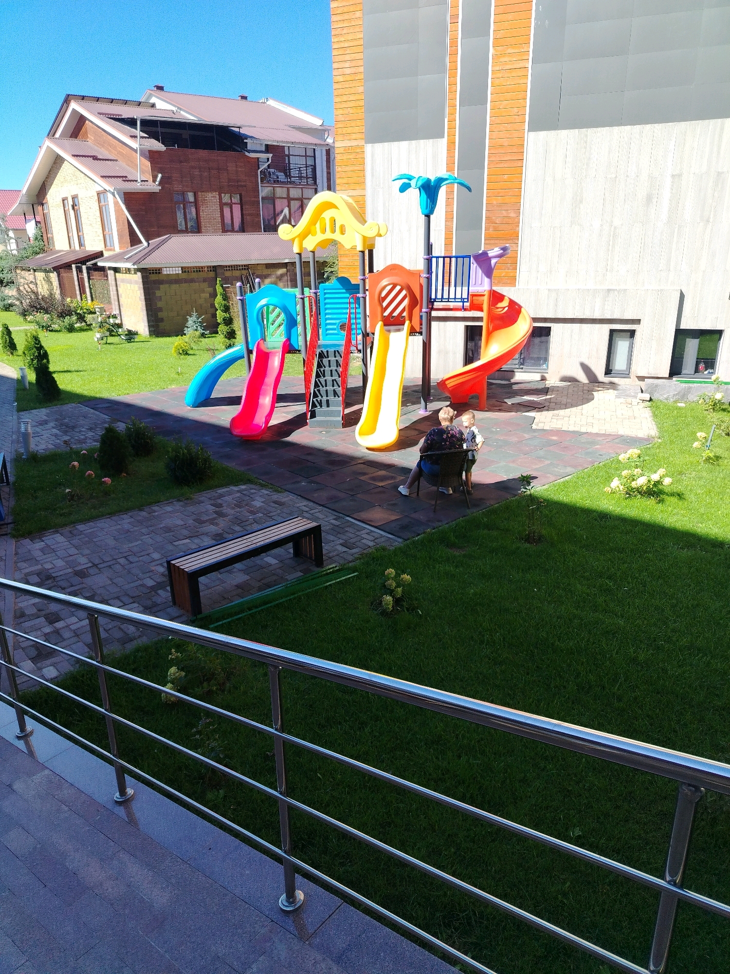 The image depicts a contemporary children's playground adjacent to a multi-story building with a modern facade featuring brick and wood elements. The playground is equipped with colorful slides, a climbing structure, and a blue archway. In the foreground, a metal railing overlooks a manicured lawn and a paved walkway with a wooden bench. Two individuals, an adult and a child, are seated on a chair near the playground, interacting with each other. The background reveals a residential building with a brown roof and a clear blue sky. The scene suggests a well-maintained outdoor recreational area.