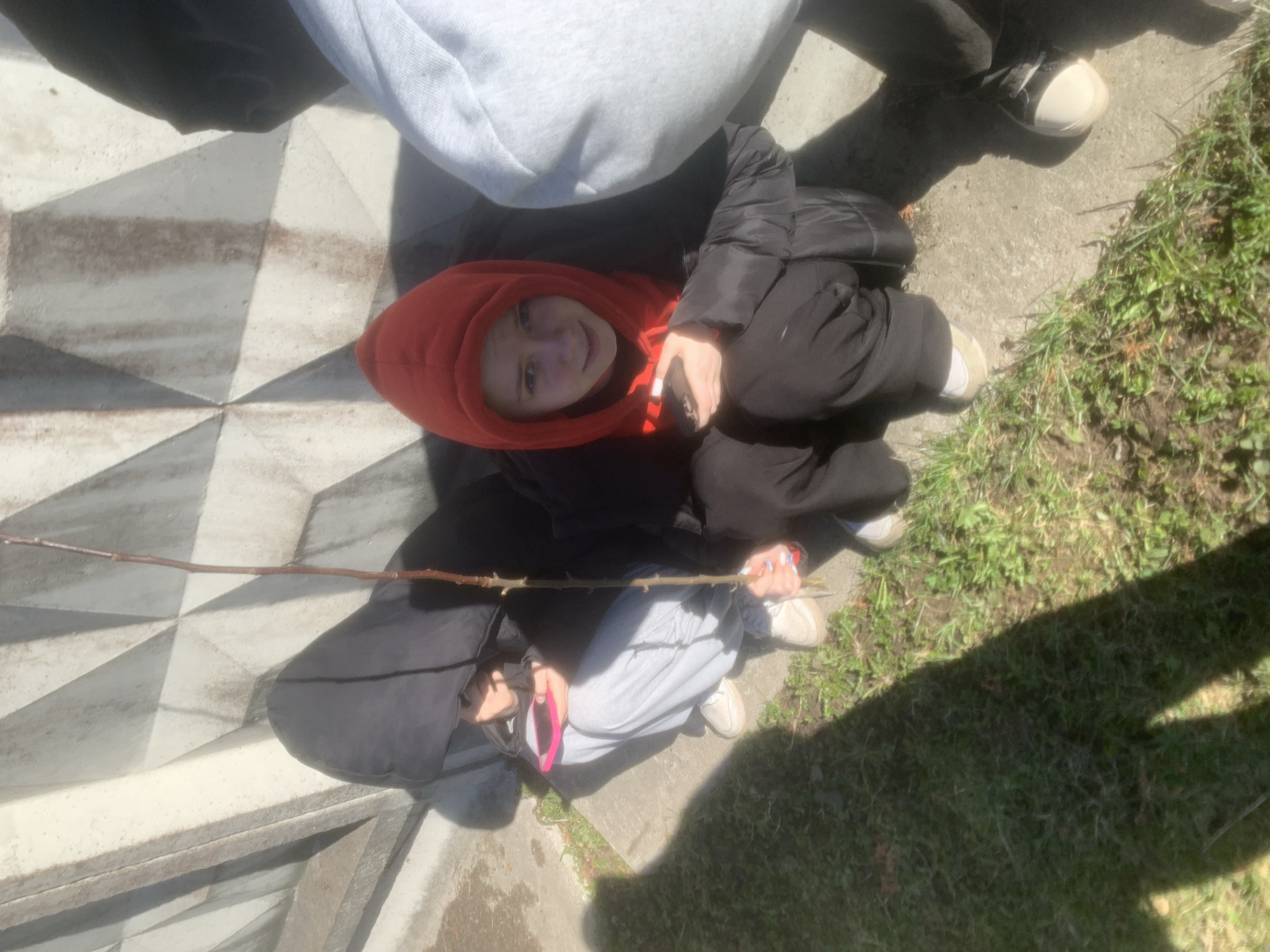 Two individuals are visible in this slightly skewed overhead shot, possibly taken in Saint Petersburg, Russia, given the geometric paving stones reminiscent of the city's architecture. One person wears an orange hooded jacket and is smiling. The other person has on a black hooded jacket and is holding a pink mobile phone. They are both holding a long stick with thorns. The surrounding environment features concrete structures, and patches of grass. One individual is wearing black and white shoes, the other is wearing plain white shoes. The lighting suggests it is daytime.