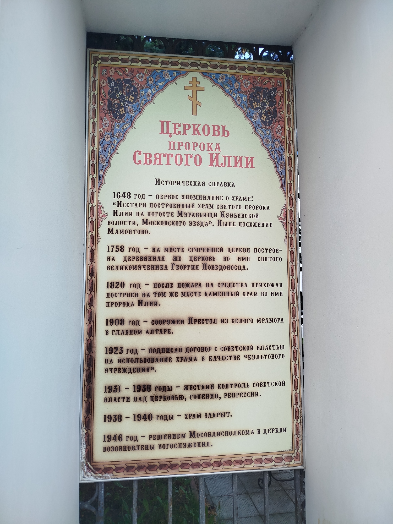 The image shows a sign with information about the "Church of the Holy Prophet Elijah". The sign provides a historical reference: 1648 - the first mention of the temple, 1758 - on the site of the burned church, a wooden church was built, 1820 - after the fire, at the expense of parishioners, a stone church was built, 1908 - a throne of white marble was built, 1923 - an agreement was signed with the Soviet government, 1931-1938 - Soviet control over the church, persecution, repression, 1938-1940 - the temple was closed, 1946 - the worship services were resumed. The sign is in Russian.
The inscription mentions the village of Mamontovo.