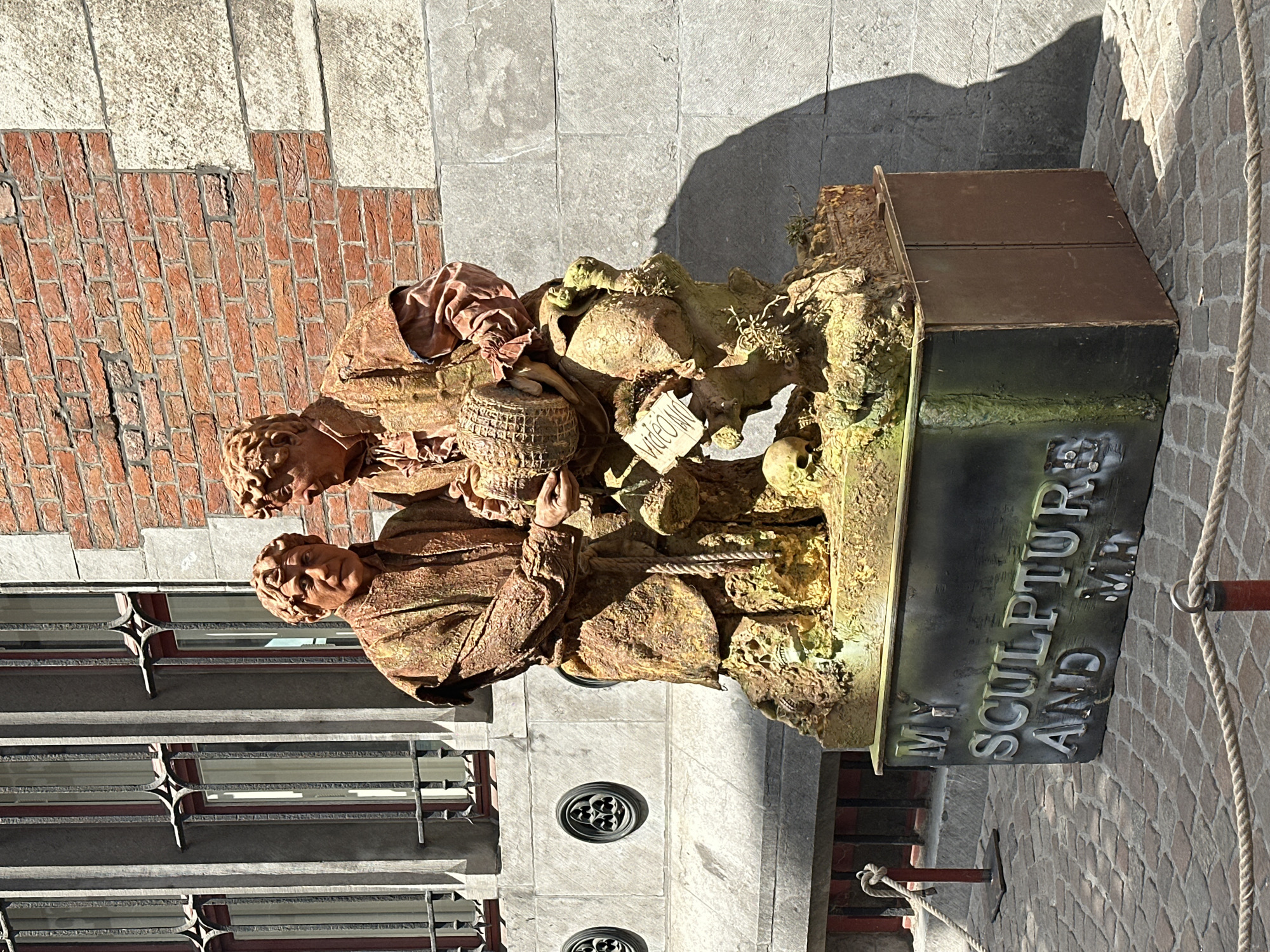 This media file captures a weathered sculpture of two figures, possibly beggars or laborers, huddled together. The sculpture is situated outdoors, with a backdrop of textured brick and stone architecture, hinting at an old-world European setting, possibly Bruges, Belgium. The figures are rendered in earthy tones, with fabric drapery that appears aged and worn. One figure holds a basket, while the other's hands are clasped. The sculpture itself is part of a larger installation, which includes a base with a dark, metallic plaque. The plaque is inscribed with the raised lettering: "MY SCULPTURE AND MR." The scene is illuminated by natural sunlight, casting distinct shadows, suggesting it is daytime with clear weather. The overall mood conveyed is one of hardship and resilience, perhaps reflecting historical or social commentary. The foreground and background elements of brickwork and stonework contribute to the historical ambiance.