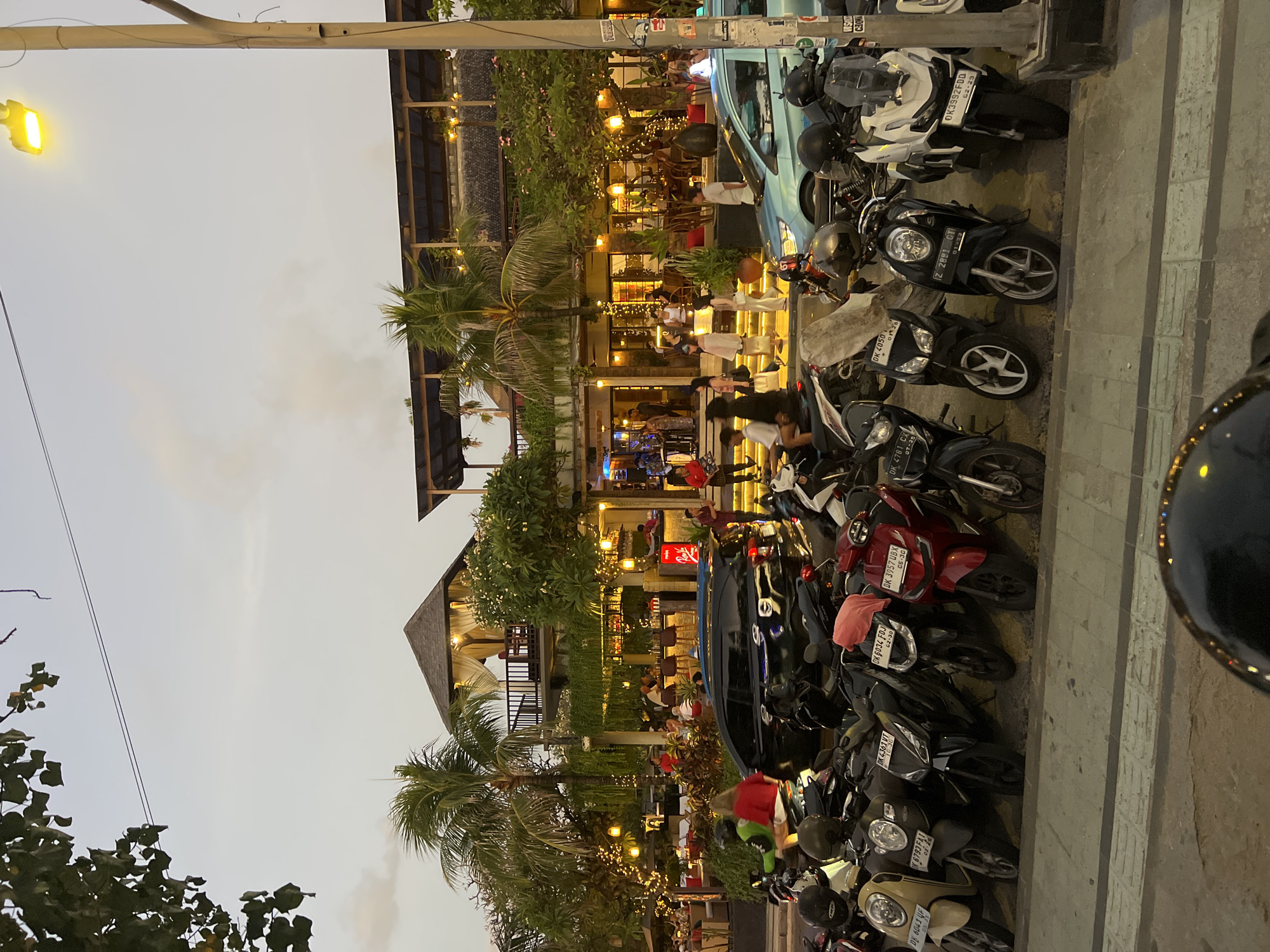 In Kuta, Indonesia, this image captures a bustling street scene in the evening. A two-story building, possibly a restaurant or bar, with an open-air upper level, is illuminated by numerous string lights. Palm trees and lush foliage surround the establishment. A prominent street lamp with a bright yellow bulb illuminates the sky. In the foreground, a large number of parked motorcycles are lined up along the sidewalk. Several people are visible walking and dining within the building, and some individuals are on motorcycles on the street. A black car is also parked in the street. The sky is a pale orange and gray, indicating dusk.