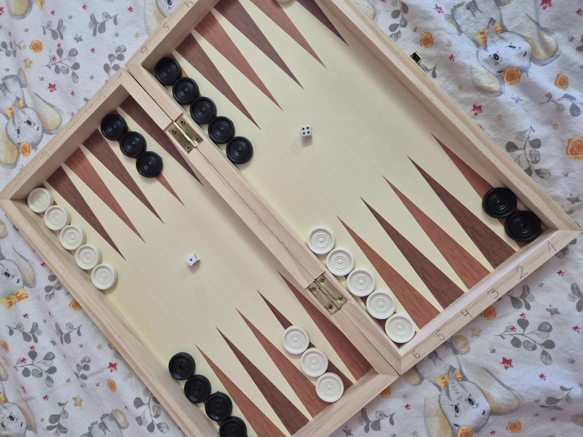 The image shows a wooden backgammon board with black and white checkers, set up on a white cloth with a whimsical pattern of cartoon elephants, leaves, and flowers. Two dice are visible on the board; one shows a '3' and the other shows a '4'. The board is open, revealing its playing surface, which is divided into triangular points of alternating dark brown and light wood colors. Some checkers are arranged on the points. The scene is photographed from directly above, with no people or identifiable landmarks present. The setting is indoors, with the cloth appearing to be a blanket or sheet.
