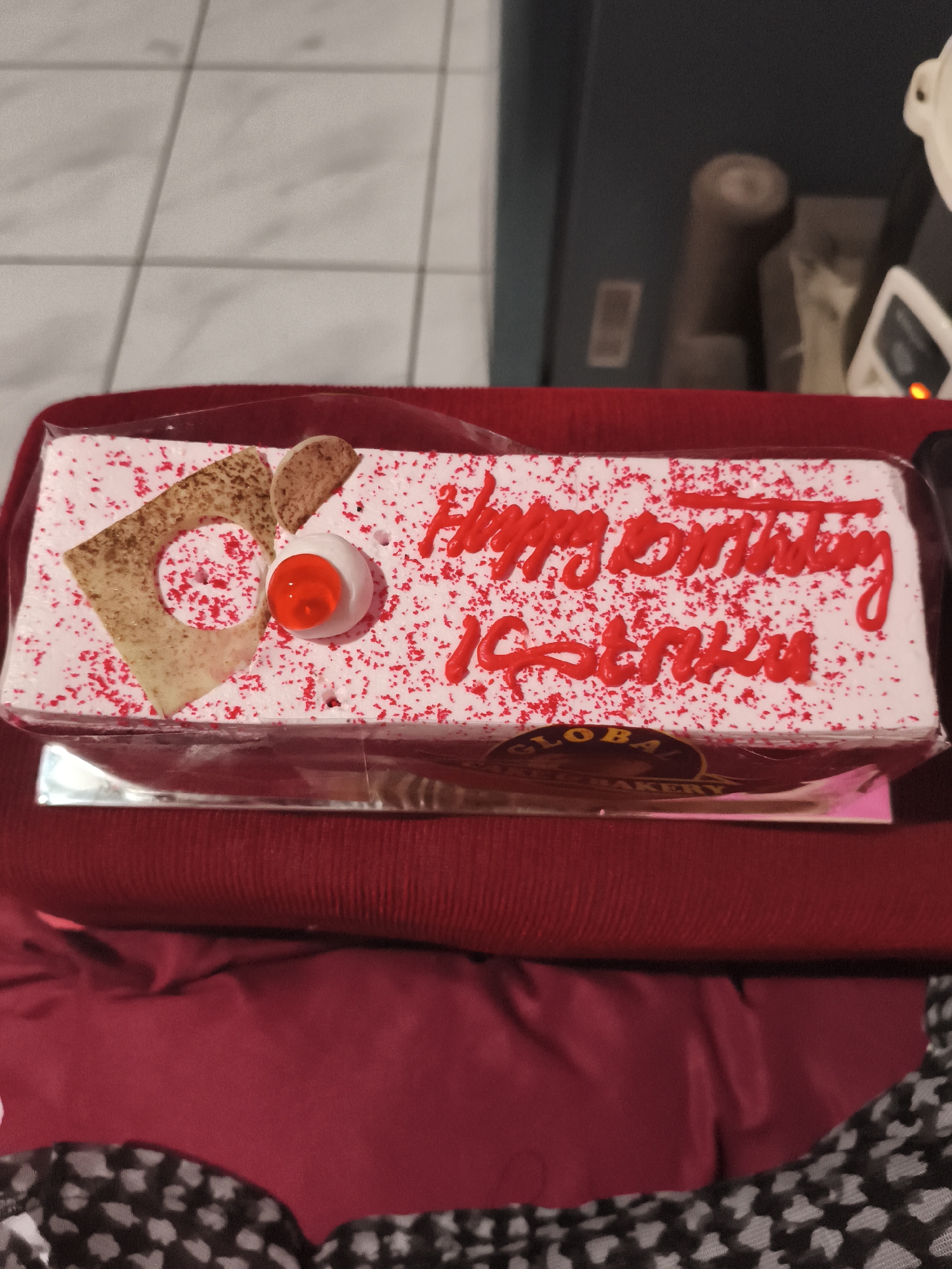 A rectangular cake with white frosting and red sprinkles sits on a red cloth. The cake has "Happy Birthday" written in red icing on top. Below that, there is another line of red icing that appears to say "Chiku". The cake is decorated with a piece of brown pastry shaped like a triangle with a hole in it, a small semi-circular piece of pastry, and a red candy. A label for "GLOBAL CAKE BAKERY" is visible on the side of the cake box. The background shows a tiled floor and some blurry objects. The image does not provide any information about the city of Depok, Indonesia.
