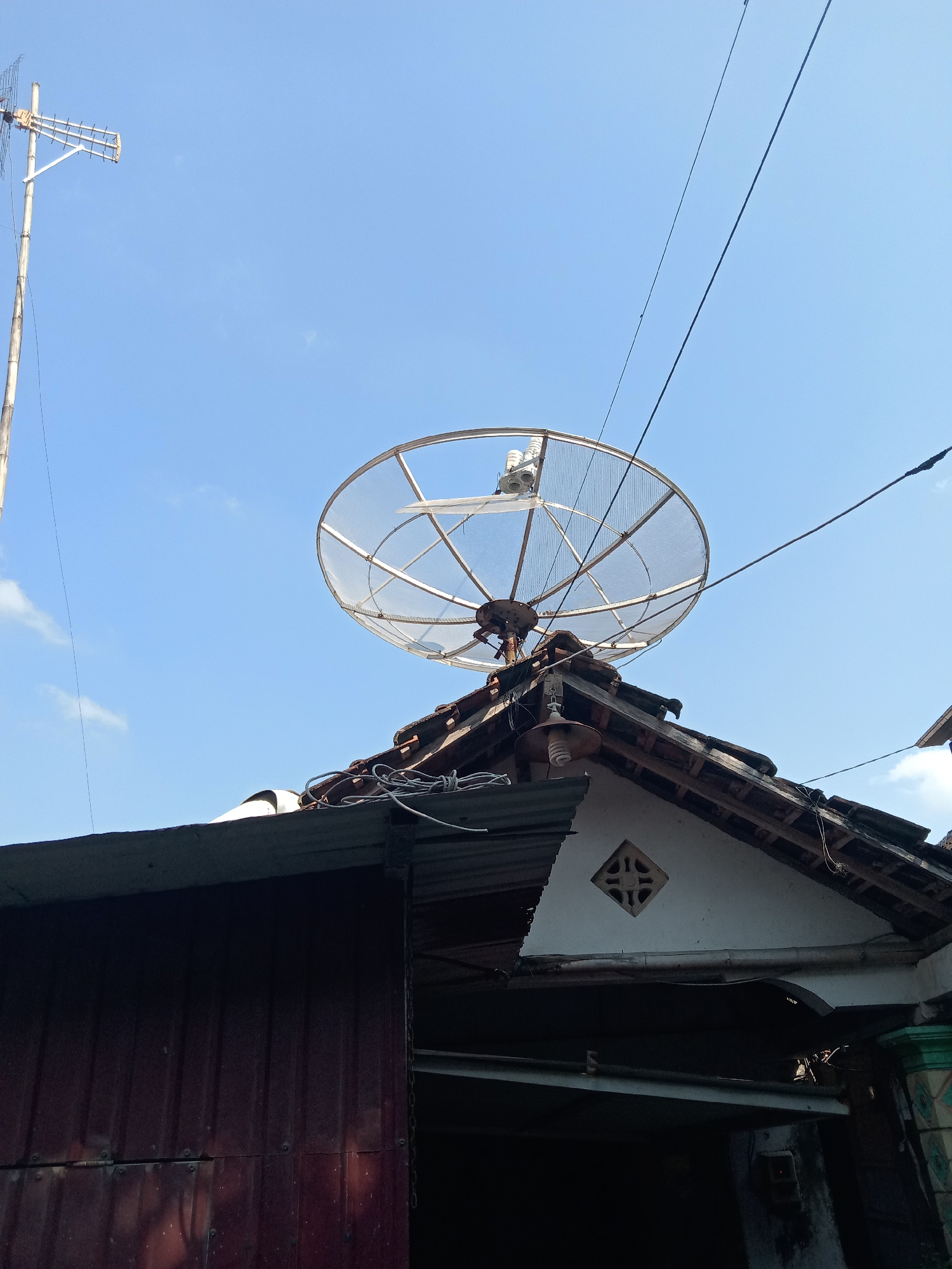 A satellite dish is mounted on the roof of a residential building, possibly in the city of Sugihwaras, Indonesia. The dish is a parabolic reflector with a white mesh surface, supported by a rusted metal arm. It is positioned above a tiled roof, with wires and cables running around it. To the left, a tall antenna on a pole stands against a clear blue sky. The building's roof has visible wooden supports and appears to be covered with dark tiles. In the foreground, a portion of a dark red corrugated metal wall is visible, suggesting it is part of the same or an adjacent structure. The overall scene depicts a common urban or semi-urban rooftop environment with telecommunications equipment.
