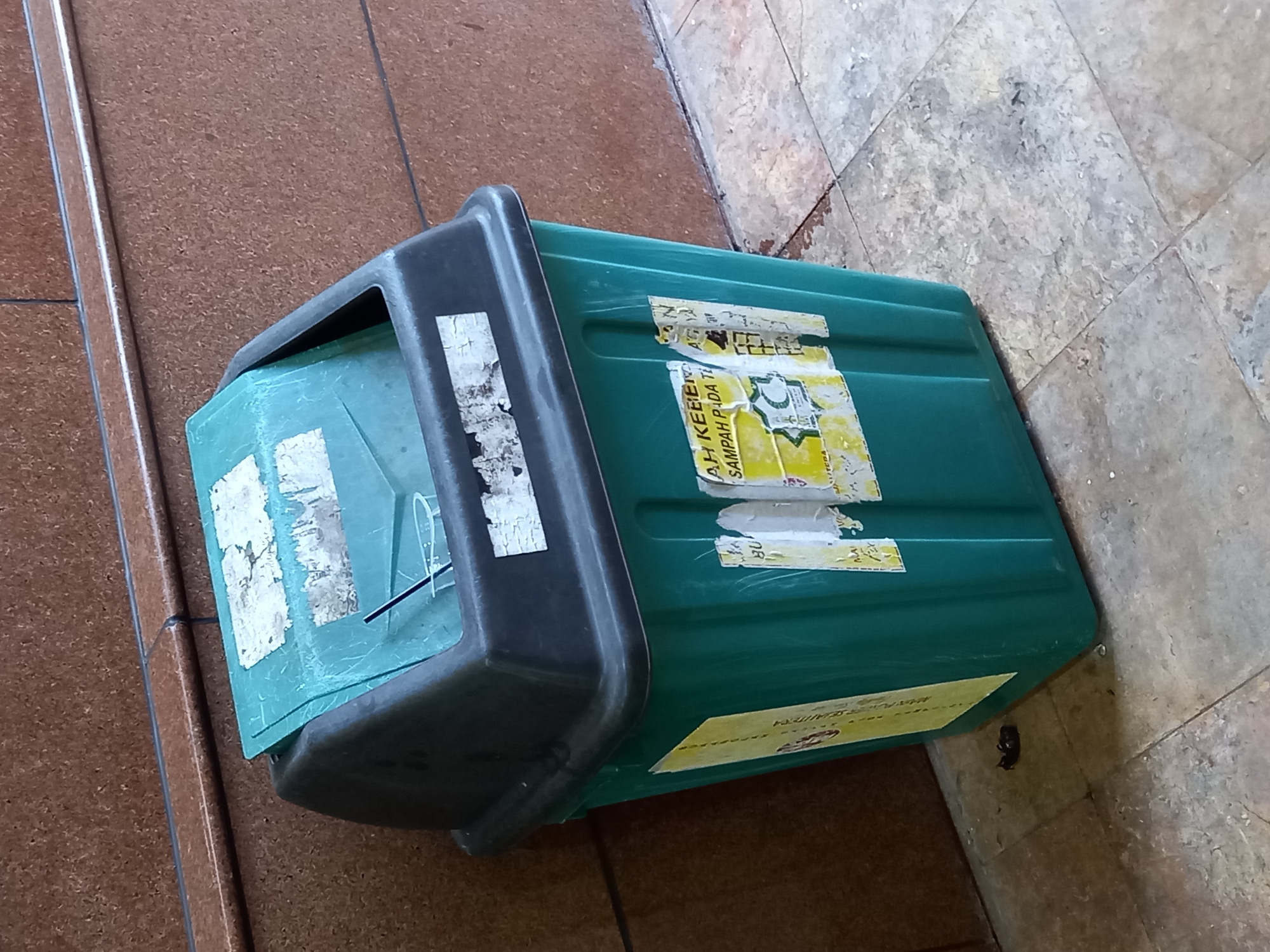 A green plastic trash can with a black lid is situated on a tiled floor in front of a brown textured wall. The trash can has a hinged lid that is partially open, revealing a clear plastic cup and a black straw inside. Several torn and faded labels are affixed to the trash can. On the left side of the trash can, a long yellow sticker with Indonesian text is visible. On the front of the trash can, there are multiple labels, including one with Indonesian text that appears to say "AH KEBER," and another with a circular emblem. A small, dark object, possibly a piece of debris or an insect, rests on the floor to the left of the trash can. The environment appears to be an outdoor or semi-outdoor public space, possibly in a commercial area or near a building. The overall condition of the trash can and its surroundings suggests a moderately used urban setting. Based on the Indonesian text on the labels, the location is likely in Indonesia. Without further context or identifying landmarks, pinpointing the exact location within Samarinda, Indonesia is not possible. However, the scene depicts a common urban element in a public space.