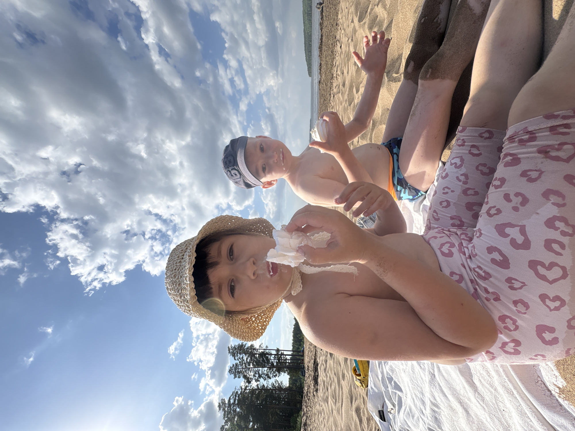 The photograph shows two children on a sandy beach. One child is wearing a straw hat with a white lace chin strap and is holding what appears to be a wad of paper or cloth. Some of the material is stuck to their mouth. The second child is in the background with a swim cap, holding a round object, possibly a ball or toy. The children are shirtless and appear to be enjoying a snack or playing with something found on the beach. The location is presumed to be in Chaikovsky, Russia, based on the request. A line of trees borders the beach in the background. The sky is filled with puffy clouds.