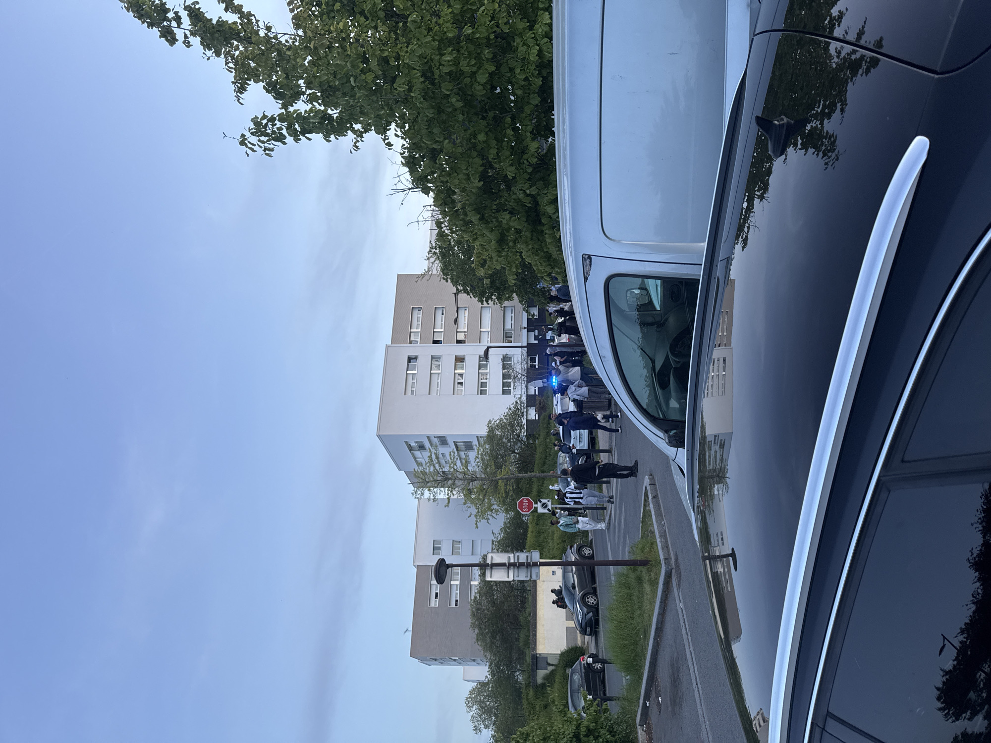 The image captures a daytime scene in a residential urban area, likely Les Ulis, France. The perspective is from ground level, looking across a street towards apartment buildings. The sky is a clear, light blue with minimal cloud cover, suggesting pleasant weather.
In the immediate foreground on the right, part of a black car is visible, showing its glossy roof and reflective side windows, mirroring some of the distant trees and buildings. To its left, a white van is parked, partially obscuring the view of the street beyond.
The mid-ground reveals a gathering of people, mostly young individuals, assembled on the side of the road. Among them, a vehicle with prominent blue flashing lights, likely an emergency or police vehicle, is noticeable, indicating an ongoing incident or official presence. Further down the street, a dark-colored car is overturned on its side, suggesting an accident. Several other cars are parked along the street.
Street furniture includes a red hexagonal stop sign mounted on a pole, clearly displaying the word "STOP". Other street signs and a lamppost are also visible. Green trees with dense foliage are interspersed throughout the scene, and grassy strips line the paved road.
In the background, several modern, multi-story apartment buildings with light-colored facades (white and beige) and numerous windows and balconies define the urban setting. The overall atmosphere suggests a casual but potentially eventful gathering within a typical European residential neighborhood.