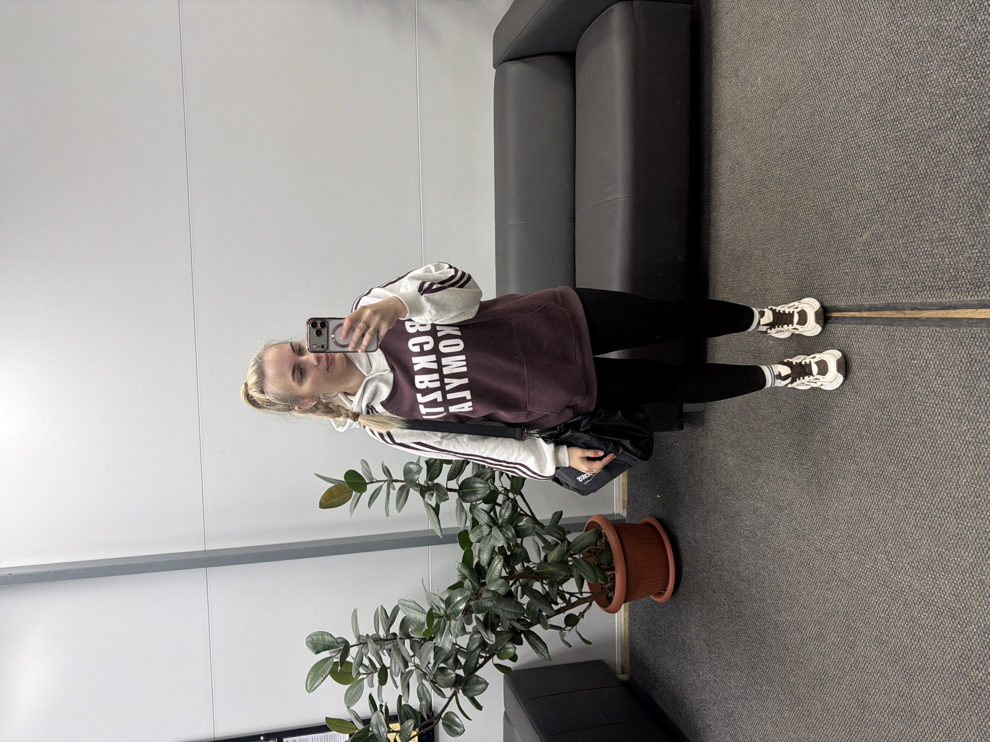 A young woman is captured taking a mirror selfie in an indoor setting in Chaikovsky, Russia. She stands, holding a smartphone with a clear case and magnetic ring in her right hand to photograph her reflection. Her blonde hair is styled in a braid.
She is dressed in casual attire, consisting of a maroon and white hooded sweatshirt with white stripes on the sleeves, black leggings, and white and brown athletic shoes. The sweatshirt features visible Cyrillic text, reversed due to the mirror, which reads "КОМАНДА" (KOMANDA, meaning "Team") above "ВСКРЫЛ" (VSKRYL, meaning "revealed" or "opened"). A black shoulder bag hangs over her left shoulder.
The background shows a contemporary interior with white walls, a grey horizontal strip, and a dark grey textured carpeted floor. To the left, a large potted plant with dark green leaves sits in a brown pot. To the right, a dark grey or black two-seater sofa is visible. The scene suggests a public or commercial indoor space, possibly a waiting area, lobby, or office hallway. The lighting is even, typical of indoor artificial illumination. The woman maintains a neutral expression for the selfie.