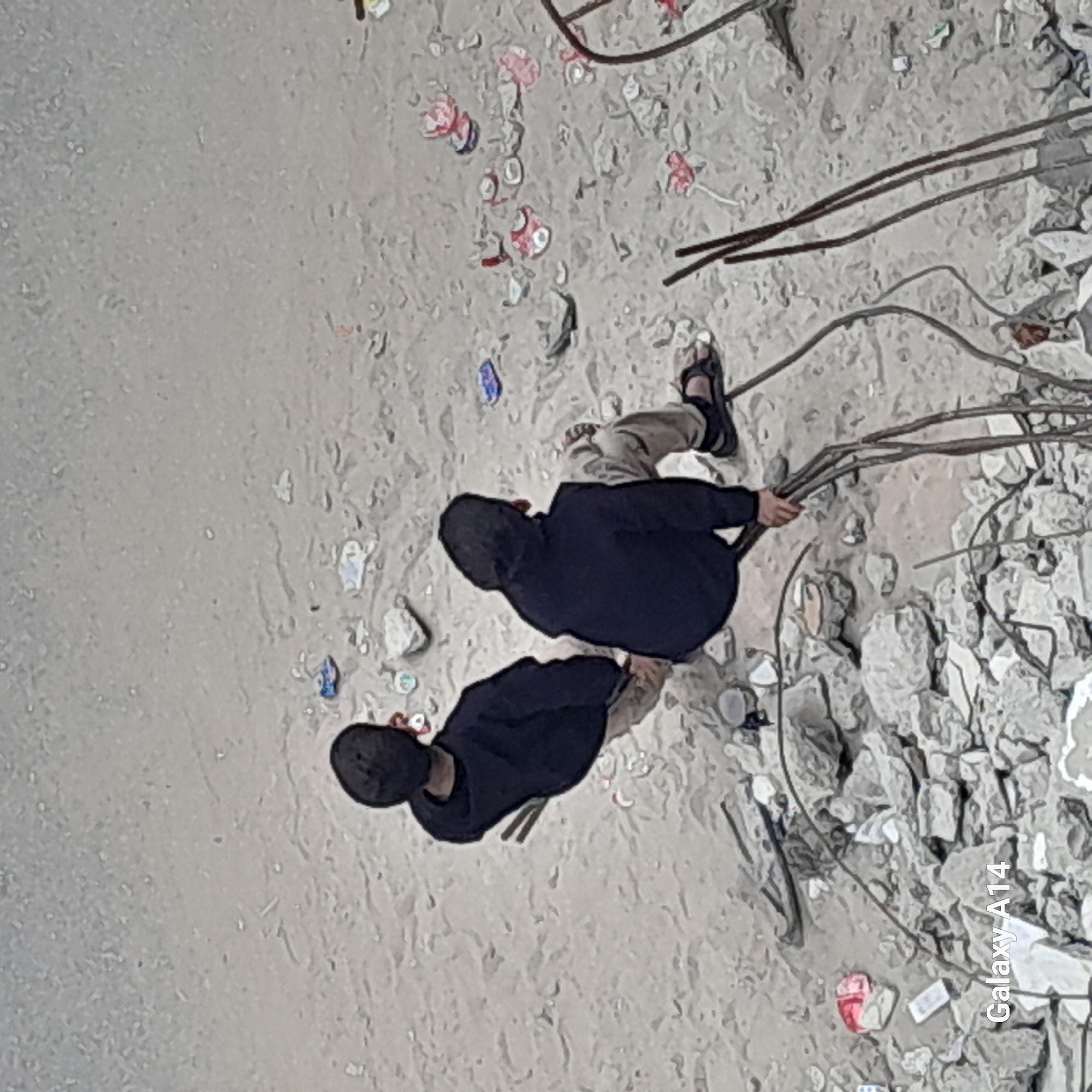 The image shows two individuals, both wearing black hooded tops and light-colored pants, standing among debris and rebar. One individual is holding a bundle of rebar. The environment includes rubble, litter, and an expanse of what appears to be sand or concrete. The location is labeled "Galaxy A14", and is in the city of Unknown, Unknown.