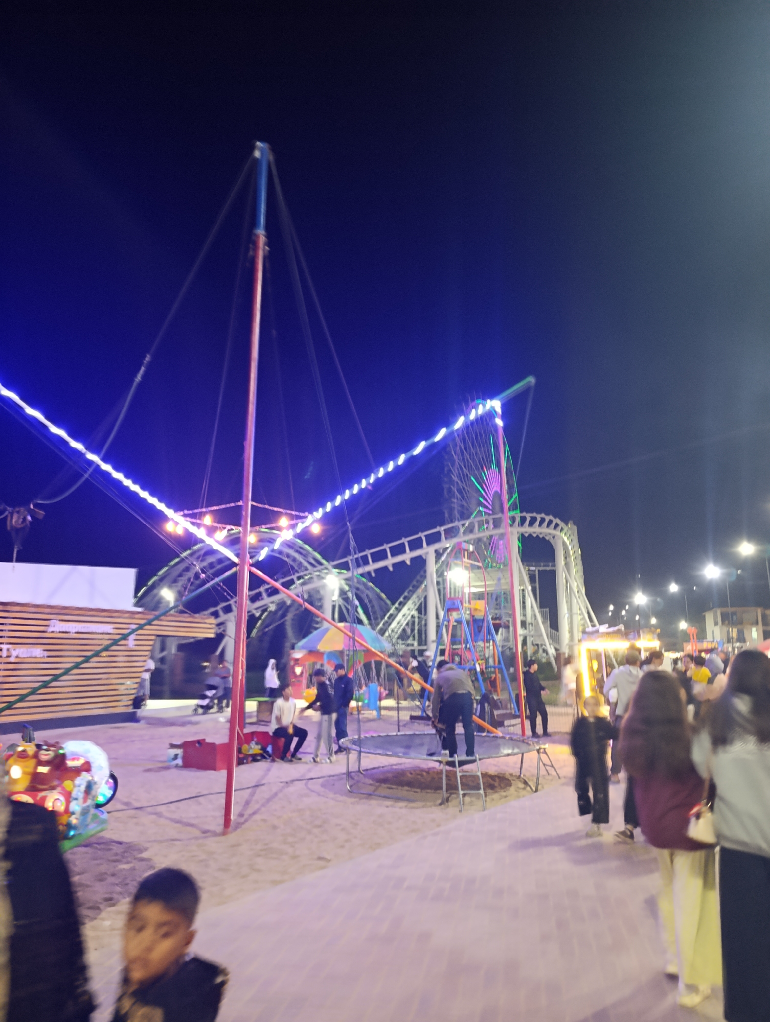 This image depicts an outdoor amusement park at night, possibly located in Bosteri, Kyrgyzstan, as indicated by the prompt. The scene is illuminated by artificial lights, including strings of blue and yellow lights decorating amusement rides. In the background, a white roller coaster track is visible, along with a Ferris wheel adorned with purple and green lights. Several people are present in the park, some standing and observing, while others appear to be interacting with the rides. One individual is standing on a trampoline, and a child is in the foreground looking towards the camera. The ground is paved with light-colored bricks. The overall atmosphere suggests a lively evening at a recreational facility.