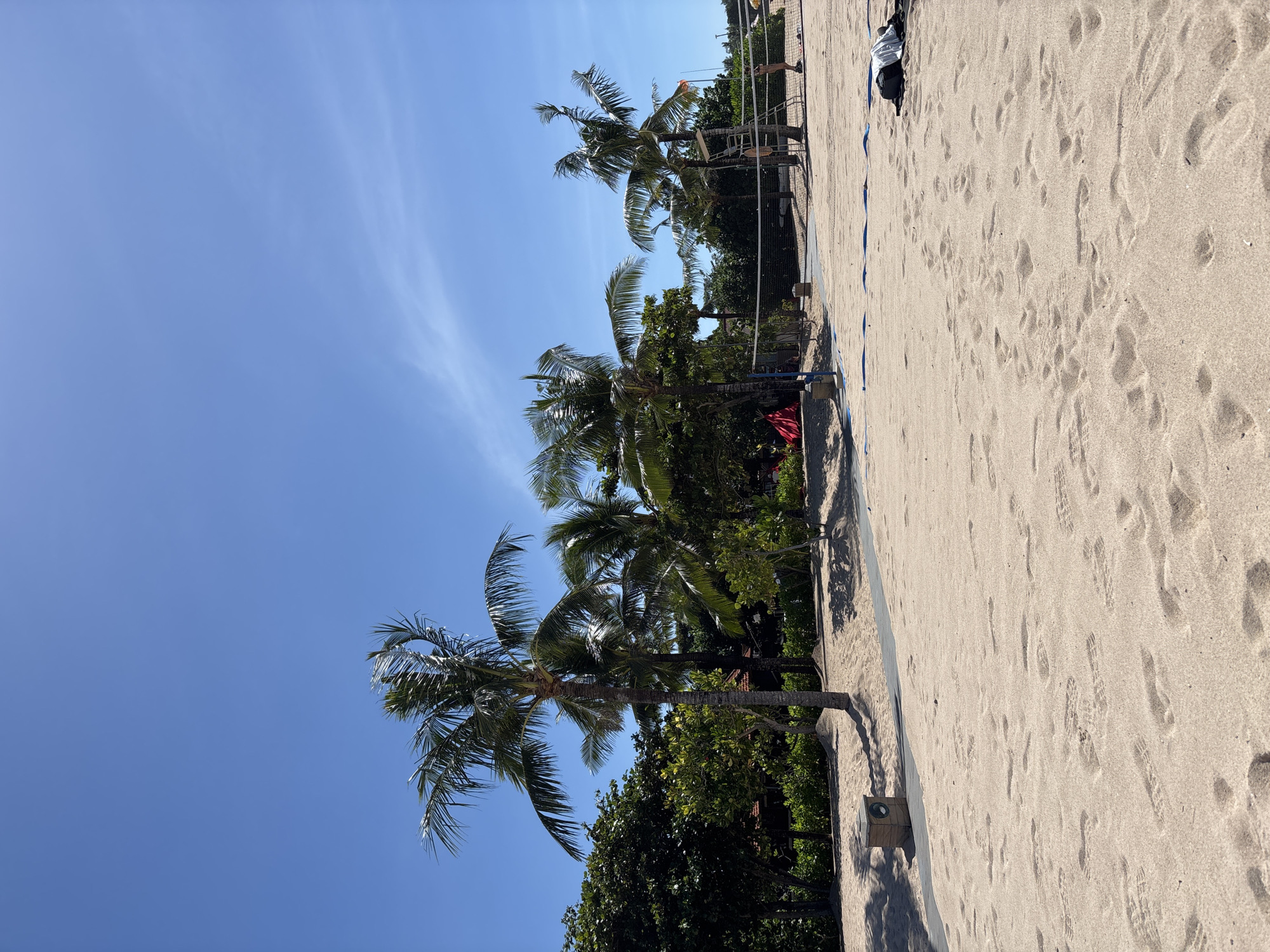 The image shows a beach scene in Kuta Selatan, Indonesia, under a clear blue sky. A line of palm trees is visible along the edge of the beach. A volleyball net is set up on the sand. The sandy beach shows numerous footprints. People can be seen near the volleyball net in the background.