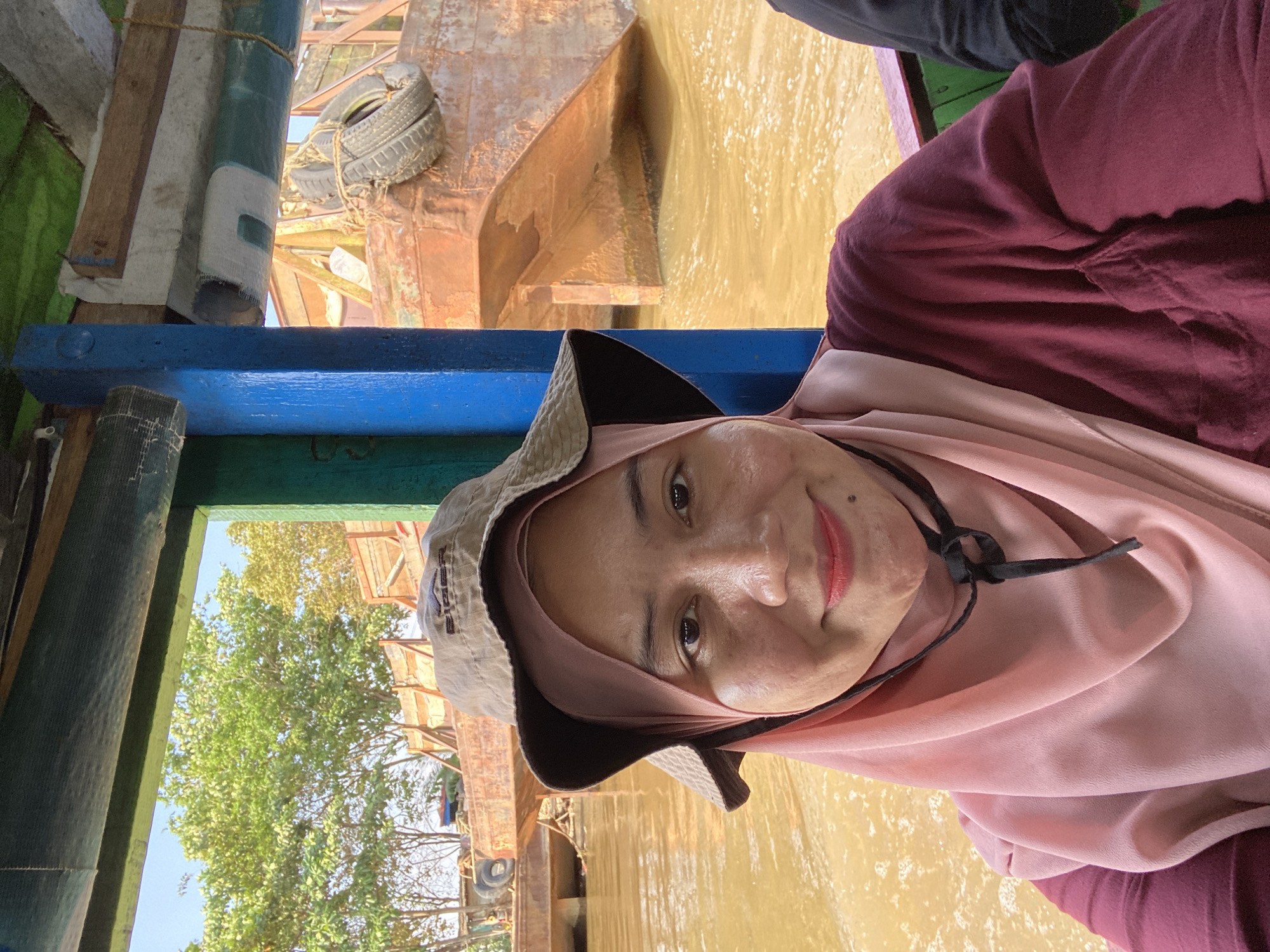 The image shows a woman wearing a pink hijab and a maroon shirt, with a tan bucket hat covering her head. She is on a boat on a river. In the background, there are other boats and some trees. The water is brown, indicating it is likely a muddy river. The woman is smiling and looking at the camera, taking a selfie. Based on the context provided, the location is the city of Banjarmasin, Indonesia, known for its many rivers and canals, making boat travel a common mode of transportation. The scene depicts a typical day of travel on a river in Banjarmasin.