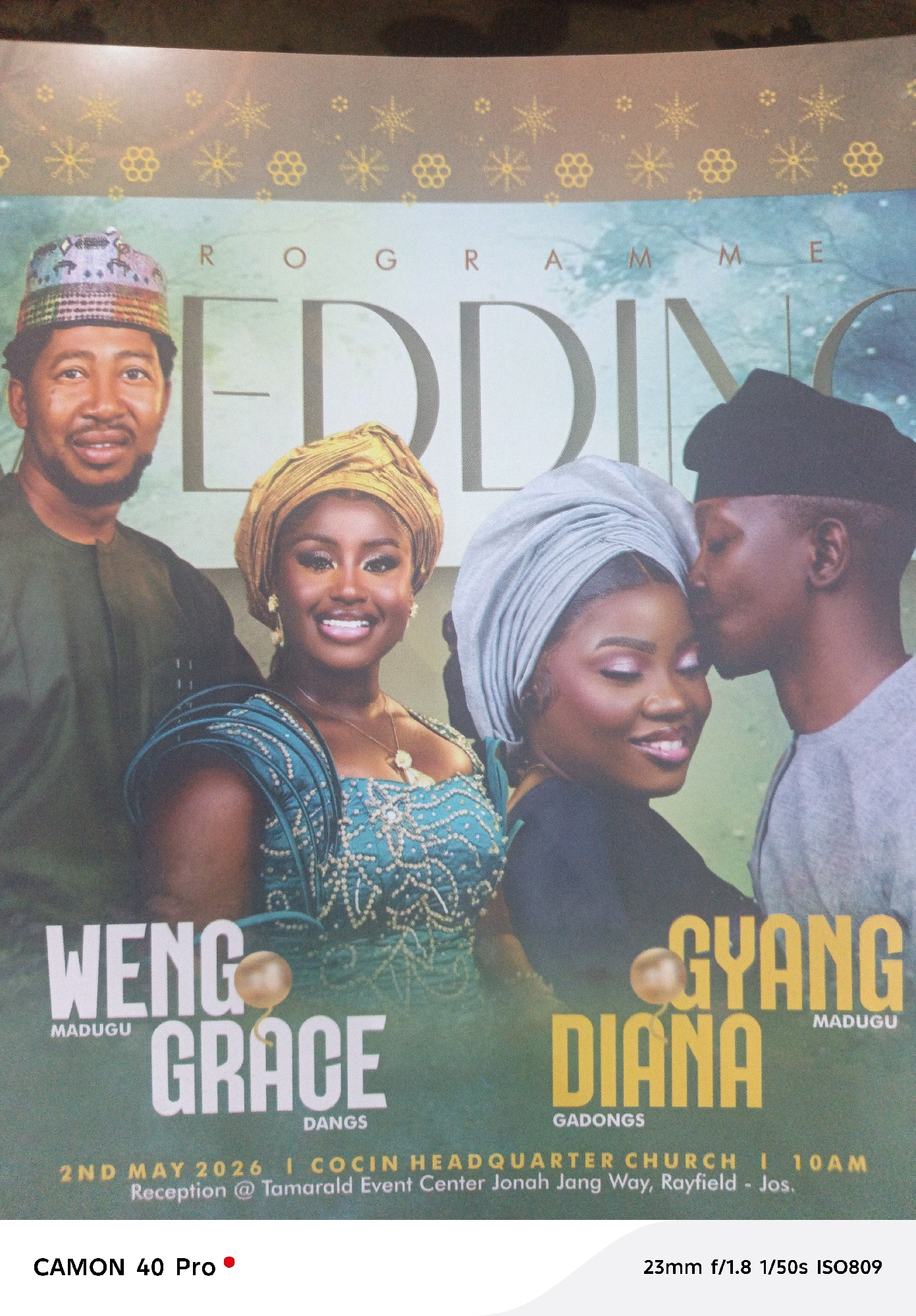 Here's a detailed description of the image:
**Subject:** The image features a wedding poster. It depicts four individuals, presumably family members of the wedding couple.
**People:**
* A man is wearing a traditional Nigerian cap, dressed in a dark green shirt.
* A woman is wearing a yellow "gele" (headwrap) and a blue and green traditional dress, smiling towards the viewer.
* Another woman is wearing a silver "gele" and is shown in profile, also smiling.
* A man is wearing a black cap and kissing the woman with the silver "gele" on the cheek.
**Text:**
* The poster has the word "WEDDING" dominating the center. Above that the word "PROGRAMME".
* On the left side, under the two people on the left are the names "WENG" above "GRACE" with "MADUGU" and "DANGS" beneath respectively.
* On the right side, under the couple on the right are the names "GYANG" above "DIANA" with "MADUGU" and "GADONGS" beneath respectively.
* At the bottom of the poster, the date is listed as "2ND MAY 2026" with "COCIN HEADQUARTER CHURCH" and "10AM"
**Setting:** The location is identified as "Reception @ Tamarald Event Center Jonah Jang Way, Rayfield - Jos."
**Environment:** The background has a pattern of gold and white floral designs. The location details indicate the city of Jos in the state of Plateau in Nigeria.
**Composition:** The image appears to be a photograph of a physical poster, as evidenced by the camera information (Canon 40 Pro, lens information).
