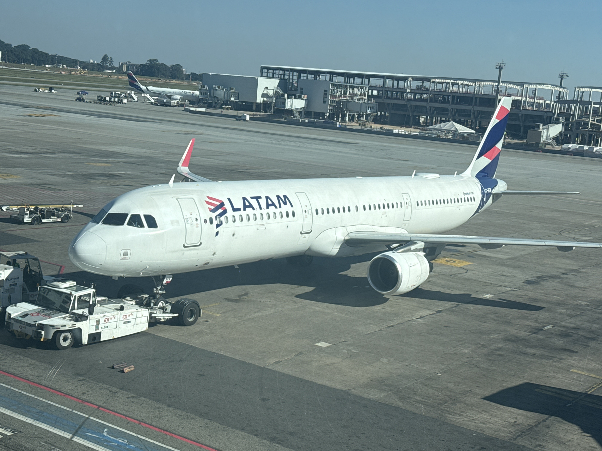 This image depicts a LATAM Airbus A321 aircraft, registration PR-XBG, parked on a concrete tarmac at São Paulo/Guarulhos – Governor André Franco Montoro International Airport (SBGR) in Guarulhos, Brazil. The aircraft, predominantly white with a blue and red LATAM livery on its fuselage and tail fin, is connected to a white WFS pushback tug (marked TL008) at its nose landing gear, indicating ground handling operations. Rows of passenger windows are visible along the fuselage, and one turbofan engine is prominent under the wing on the left side of the aircraft.
In the mid-ground, another LATAM aircraft, identifiable as an Airbus A320 family member, is parked at a gate and connected to a jet bridge. Various ground support equipment, including baggage carts and smaller service vehicles, are positioned around the distant aircraft on the tarmac. The background features a large, multi-story airport terminal structure under apparent construction or renovation, exhibiting exposed concrete and steel frameworks, with multiple jet bridges attached. A treeline borders the far perimeter of the airport, under a clear, light blue sky. Strong shadows cast by the aircraft and ground equipment suggest bright daylight conditions. No discernible people are individually visible in the scene.