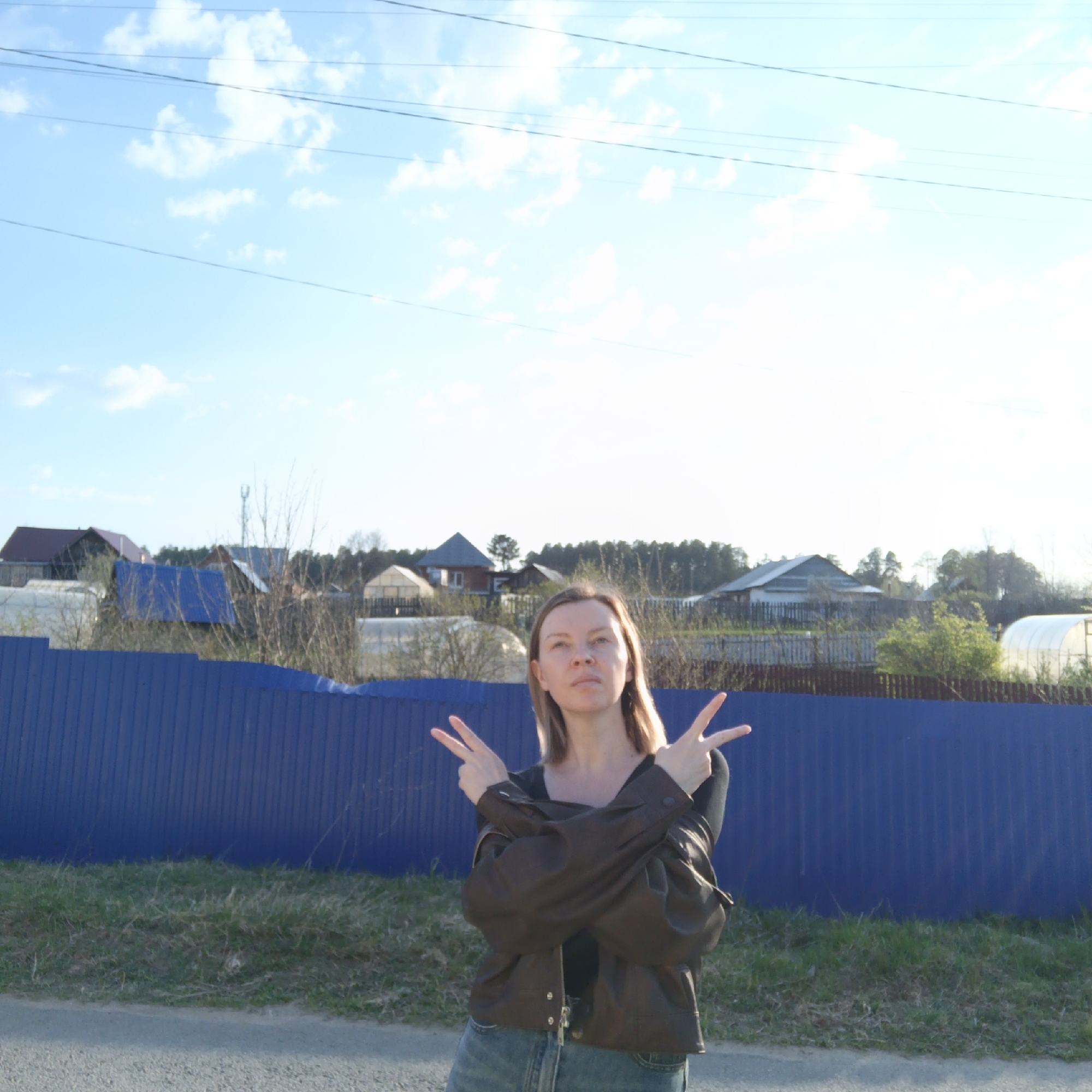 A woman stands outdoors in front of a blue metal fence and rural buildings under a bright sky with scattered clouds. She wears a brown jacket, jeans, and a dark top and holds both hands up with fingers extended, making a peace sign on both hands. The location is Chaikovsky, Russia. A rural community with small homes is visible beyond the fence.