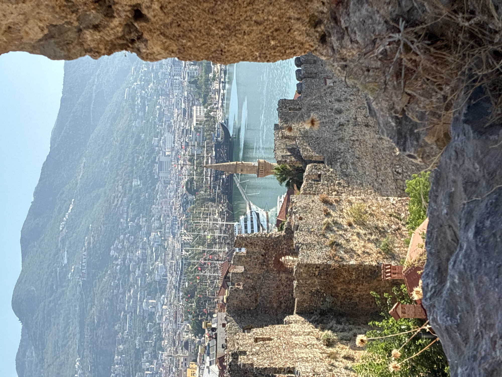 The image captures a scenic view of Alanya, Turkey, from a vantage point overlooking the harbor and the city, framed by weathered stone walls of a historical structure. In the foreground, rough-hewn stone fortifications, likely part of Alanya Castle, dominate the view, with crumbling battlements and stone blocks. To the left, glimpses of red-tiled roofs and brick chimneys of buildings nestled among greenery are visible.
The middle ground reveals a bustling harbor filled with a multitude of sailboats, their masts creating a striking silhouette against the water. A white cruise ship or large ferry is docked in the harbor, and smaller boats are also present. A tall, slender minaret with a conical roof rises from the waterfront, a distinctive architectural feature of the city. The turquoise water of the Mediterranean Sea stretches out, reflecting the clear blue sky.
In the background, the city of Alanya sprawls across the foothills, a dense collection of buildings with white and light-colored facades. Ascending the steep hillside behind the city, a prominent white sign spells out "ALANYA" in large letters, a well-known landmark. The lush green slopes of the mountain form the backdrop, disappearing into a hazy sky, suggesting a warm, sunny day. The overall impression is one of historical grandeur meeting a vibrant coastal city.