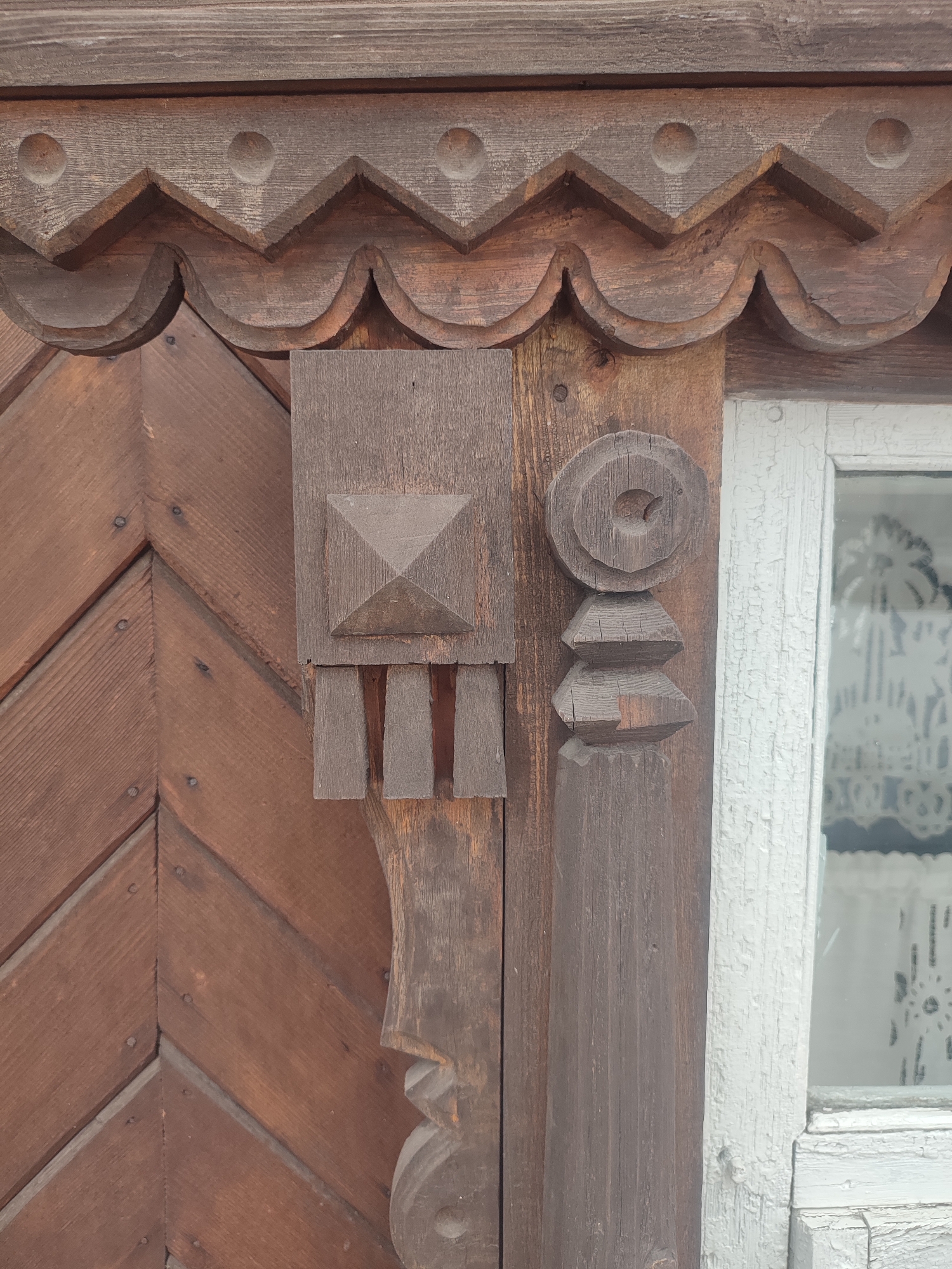The image depicts a close-up of intricately carved wooden architectural details, likely from a building in Sarapul, Russia. The wood is stained a dark brown color and shows signs of weathering. A trim along the top of the frame consists of a pattern of zig-zags and rounded scallops. Below that, a vertical structure is divided into two distinct parts. On the left, the wooden detail features a square section with a raised pyramidal shape, followed by three short, vertical bars underneath. Further down, a stylized carving resembling a face or mask is visible. To the right, adjacent to a window with a white frame and sheer curtain, another vertical wooden detail displays a circular element with a hole at the center, followed by two stacked trapezoidal shapes, and a more elaborate, carved portion near the bottom. The wooden planks to the left of the decorations are arranged in a herringbone pattern and secured with visible nails. The window, with its peeling white paint, suggests that this part of the building is old.