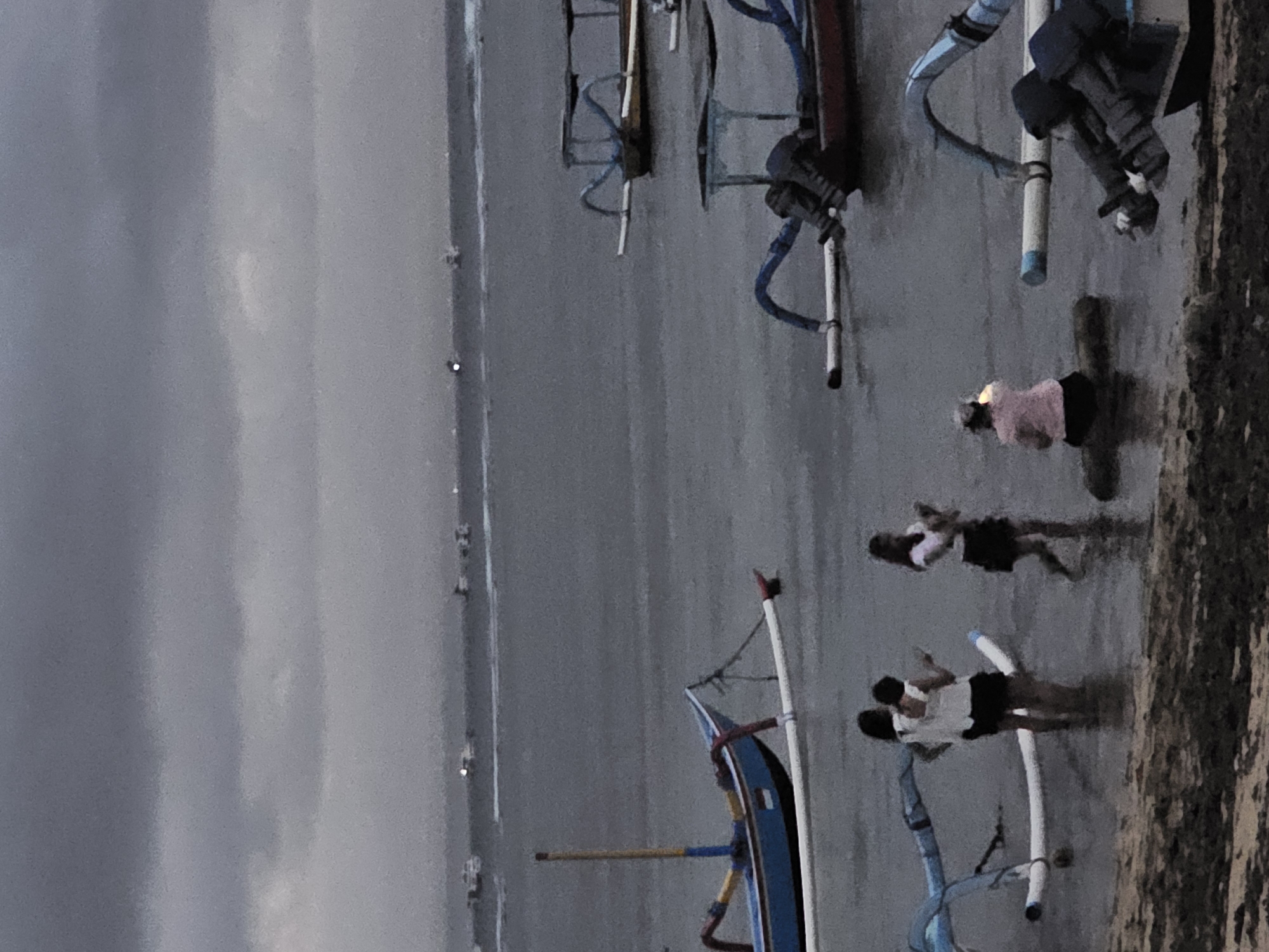 The image depicts a coastal scene in Denpasar, Indonesia, characterized by overcast skies and calm waters. Several traditional outrigger boats, known as jukungs, are moored near the shore. In the foreground, a person wearing a dark hooded jacket is seated in a boat. Further out, three individuals are standing in the shallow water. One person is wearing a pink shirt and black shorts, another is wearing a white top and black skirt, and the third is wearing a white top and black skirt. A blue and yellow jukung is anchored closer to the shore. In the distance, multiple boats are visible on the water, and a faint horizon line suggests a vast expanse of sea. The overall atmosphere is subdued, possibly due to the weather conditions.