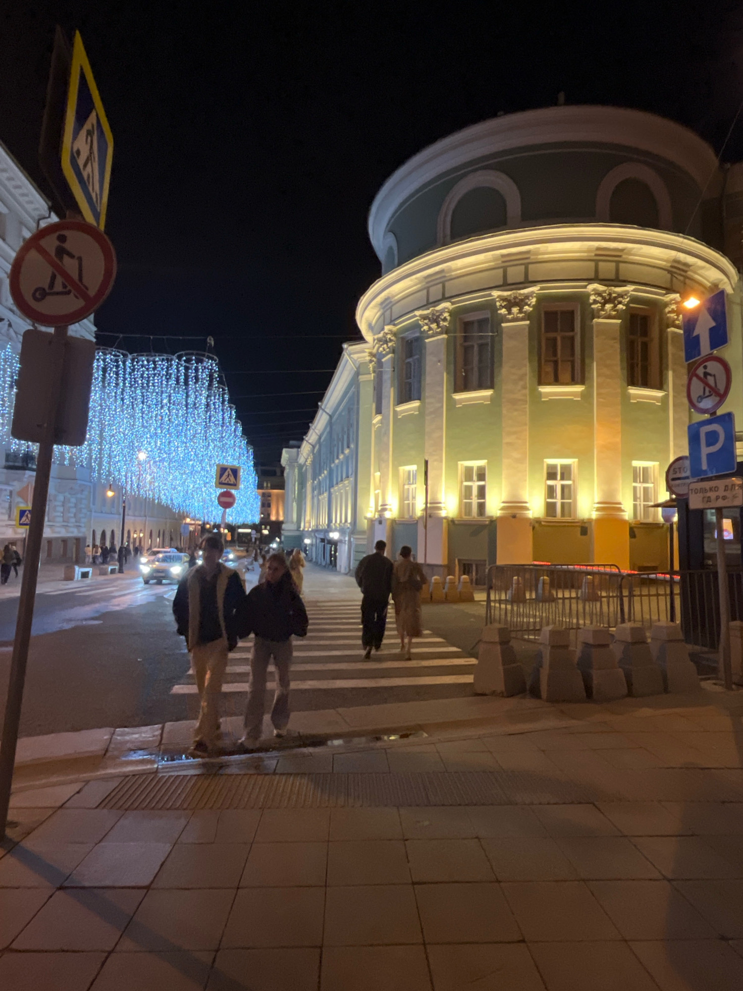 This is a nighttime, eye-level, medium shot of a street scene in Moscow, Russia, possibly on Nikolskaya Street, known for its festive illuminations. In the foreground, a man and a woman are walking across a pedestrian crosswalk, illuminated by streetlights and the ambient glow of the city. The man is wearing a dark jacket and light-colored pants, while the woman is wearing a dark jacket and light-colored pants. They are walking from left to right, facing away from the camera.
To the left of the pedestrian crossing, a street sign with a "no scooters" symbol and a diamond-shaped sign indicating a pedestrian crossing are visible. The street is lined with buildings on both sides, adorned with bright blue string lights that create a dazzling canopy over the street. The building on the right features a prominent dome and is illuminated with yellow and green lights, highlighting its architectural details. Traffic lights and other street signs are also present. Cars are visible on the street, with their headlights on, suggesting ongoing traffic. The sidewalk is paved with square tiles, and there are some temporary concrete barriers along the street. The overall atmosphere is festive and urban, typical of a well-lit city street at night.