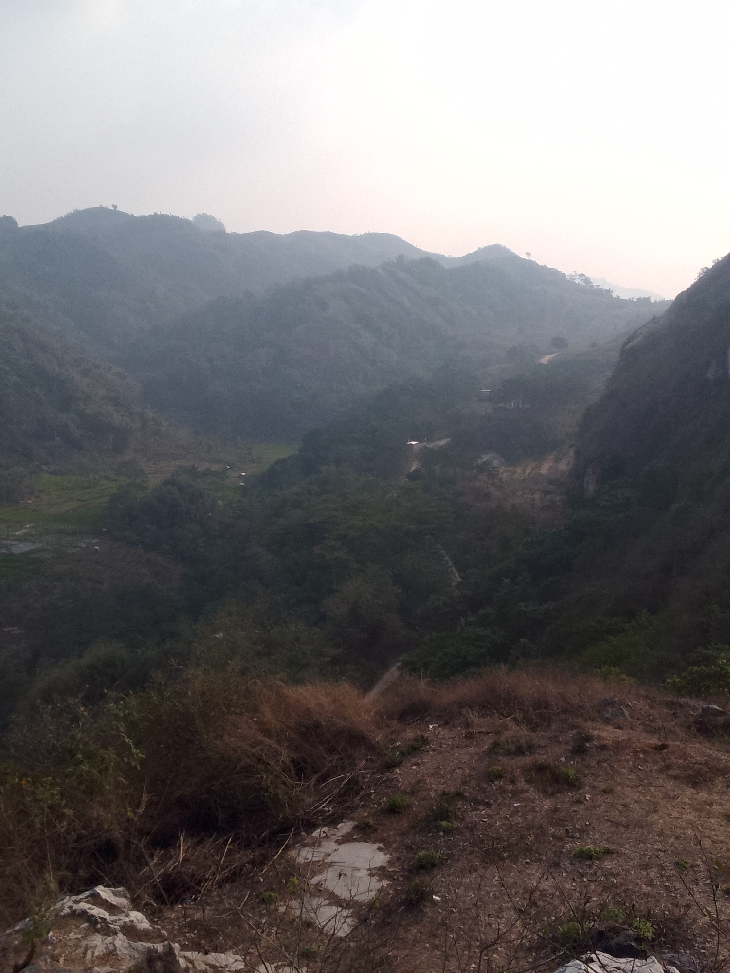 An overcast day looms over a mountainous landscape. The foreground is dominated by dry, brown vegetation and rocky terrain. Beyond this, a verdant valley unfolds, dotted with what appear to be small structures and terraced fields. The valley is framed by steep, tree-covered hills that recede into the distance, their contours softened by a hazy atmosphere. A winding dirt road is visible snaking through the landscape, hinting at human presence in this remote region. The overall impression is one of a rugged, natural environment with evidence of settlement and agriculture.