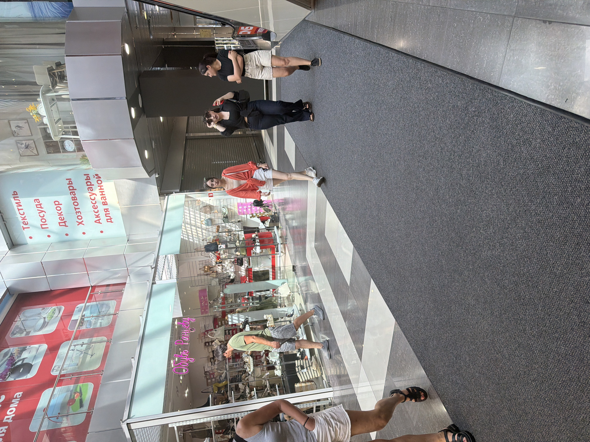 The image depicts an indoor shopping mall scene in Sochi, Russia. Three women are walking on a dark grey carpeted walkway. The woman on the left is wearing a white tank top, white shorts, and black sandals. The woman in the middle is wearing a red cropped top, white shorts, and black sandals, with sunglasses perched on her head. The woman on the right is wearing a black t-shirt, beige shorts, and black sneakers. Behind them, a man in a green t-shirt and grey shorts walks past a store with a neon sign reading "Обувь Рацевая". The store displays shoes and bags on shelves. The mall features a clean, modern aesthetic with tiled floors and large windows. The overall environment suggests a commercial retail space.