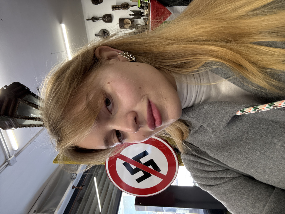 The image depicts a young adult female captured in a close-up, self-portrait style photograph, seemingly taken indoors within a commercial establishment in Cagliari, Italy, identifiable as an antique or curiosity shop.
The female subject, positioned centrally, has light-colored skin, long, straight, light brown to blonde hair with bangs, and is looking slightly towards the viewer's right with a faint smile. She is wearing pink lipstick. A silver-colored, leaf-motif ear cuff adorns her right ear. Her attire consists of a light grey, textured, open-front coat over a white turtleneck sweater. A multicolored patterned strap, likely from a bag, is visible over her right shoulder.
To the immediate left of the woman's head, a circular white sign with a thick red border and a diagonal red slash indicating prohibition is prominently displayed. Centered within this prohibitory symbol is a black swastika.
The background reveals the interior of what appears to be a retail space. The ceiling features exposed fluorescent tube lighting and a dark, multi-faceted lantern-style light fixture suspended from above. On the right, a wall displays several dark-colored, carved wooden masks or tribal art pieces. Below these, indistinct objects rest on a surface partially covered with red and white fabric. To the far left, a bright area suggests an open doorway or window leading to a blurry exterior view, with a yellow vertical support structure also visible.