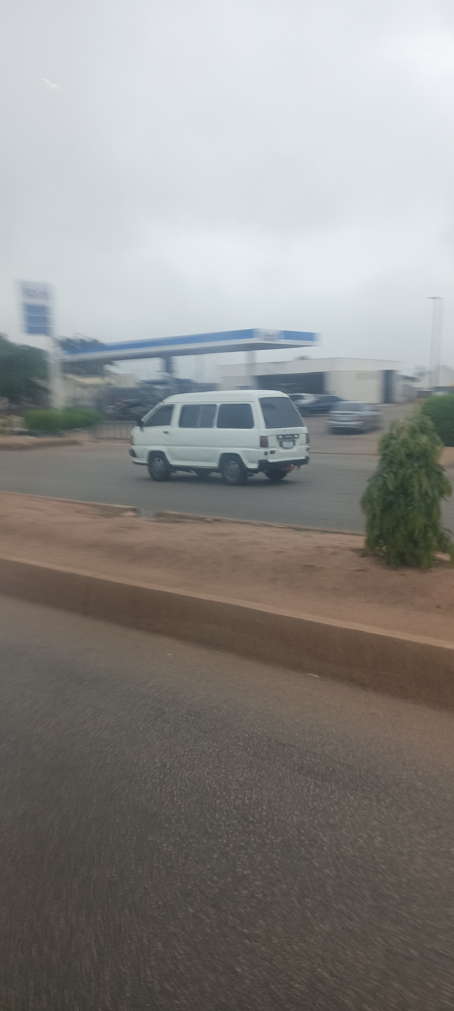 The image depicts a white van, possibly a Toyota LiteAce or a similar model, in motion on a paved road. The van is captured from a side-rear angle, driving from left to right across the frame. Its wheels appear to be turning, and the rear license plate is present but illegible due to blur.
In the background, a gas station is visible with a prominent blue and white canopy structure. A tall, blue sign, likely associated with the gas station, is also present on the left, but any text on it is too blurry to discern. Other vehicles are faintly visible and blurred in the background around the gas station area. To the right of the van, a small, green, bushy tree grows on the roadside.
The foreground consists of a dark grey paved road, a light brown raised curb, and an unpaved, sandy shoulder. The sky is overcast and hazy, indicating a cloudy daytime setting or atmospheric dust, resulting in soft, diffused lighting with no harsh shadows. The overall motion blur suggests the photograph was taken from a moving vehicle or captures the van's movement with a slower shutter speed. The scene is consistent with a roadside environment in Jos, Nigeria.