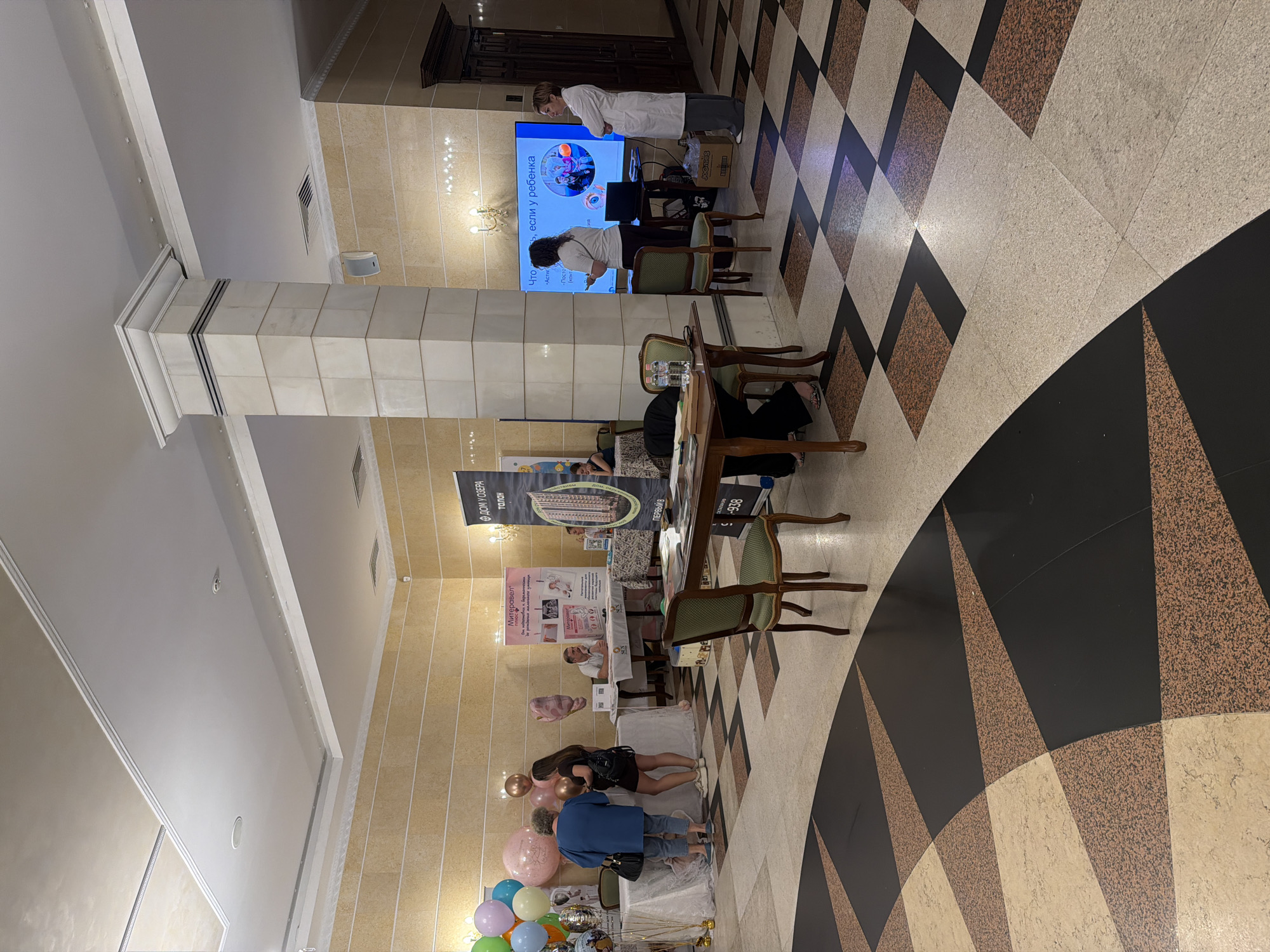 The image depicts an indoor event space, likely a conference or exhibition hall, with a patterned floor and beige tiled walls. In the foreground, a dark wood table with chairs is visible. To the right, a large screen displays text in Russian, along with images of people. Two women are standing near the screen, one in a white lab coat and the other in casual attire.
Further to the left, there are several tables set up with informational displays and balloons. Several people are gathered around these tables, interacting with each other and with the displays. One woman in a blue top and jeans is looking at a display. Another woman in a black dress is standing next to her. A man is seated behind one of the tables, seemingly engaged in conversation. Banners with Russian text are also visible, one of which appears to be advertising a real estate development.
The overall impression is that of a public event or exhibition, possibly related to healthcare, family services, or real estate, taking place in a well-lit indoor venue. The presence of balloons and informal interactions suggests a relatively relaxed atmosphere. The specific location in Khabarovsk, Russia, is not explicitly indicated within the image itself, but the Russian text on the displays and banners strongly suggests this geographical context.
