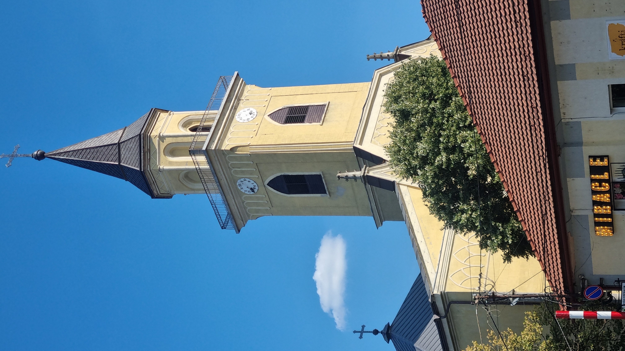 This image captures a view of the Roman Catholic Church in Berehove, Ukraine, on a clear, sunny day. The church features a tall, yellow bell tower topped with a dark steeple and a cross. The tower has arched windows and two clock faces visible. Adjacent to the bell tower is another section of the church with a dark roof and a cross. In the foreground, a building with a red tile roof is visible, with the word "MIRACLE" spelled out in illuminated letters on its facade. Trees and utility poles are also present in the scene. The sky is a bright blue with a single small white cloud.