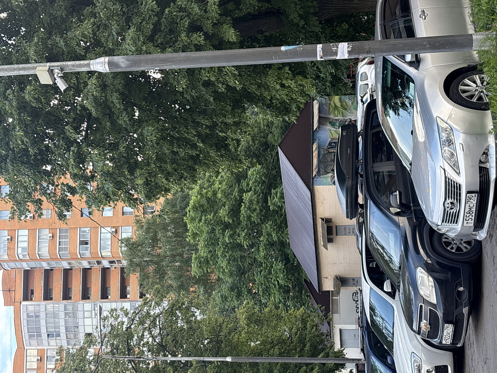 In Khimki, Russia, a line of parked cars is visible, with a streetlamp and surveillance camera in the foreground. Behind the cars, a low-slung building with a dark, sloped roof is adorned with a mural. Further back, lush green trees dominate the midground, and a multi-story brick apartment building rises in the background. The sky is overcast.