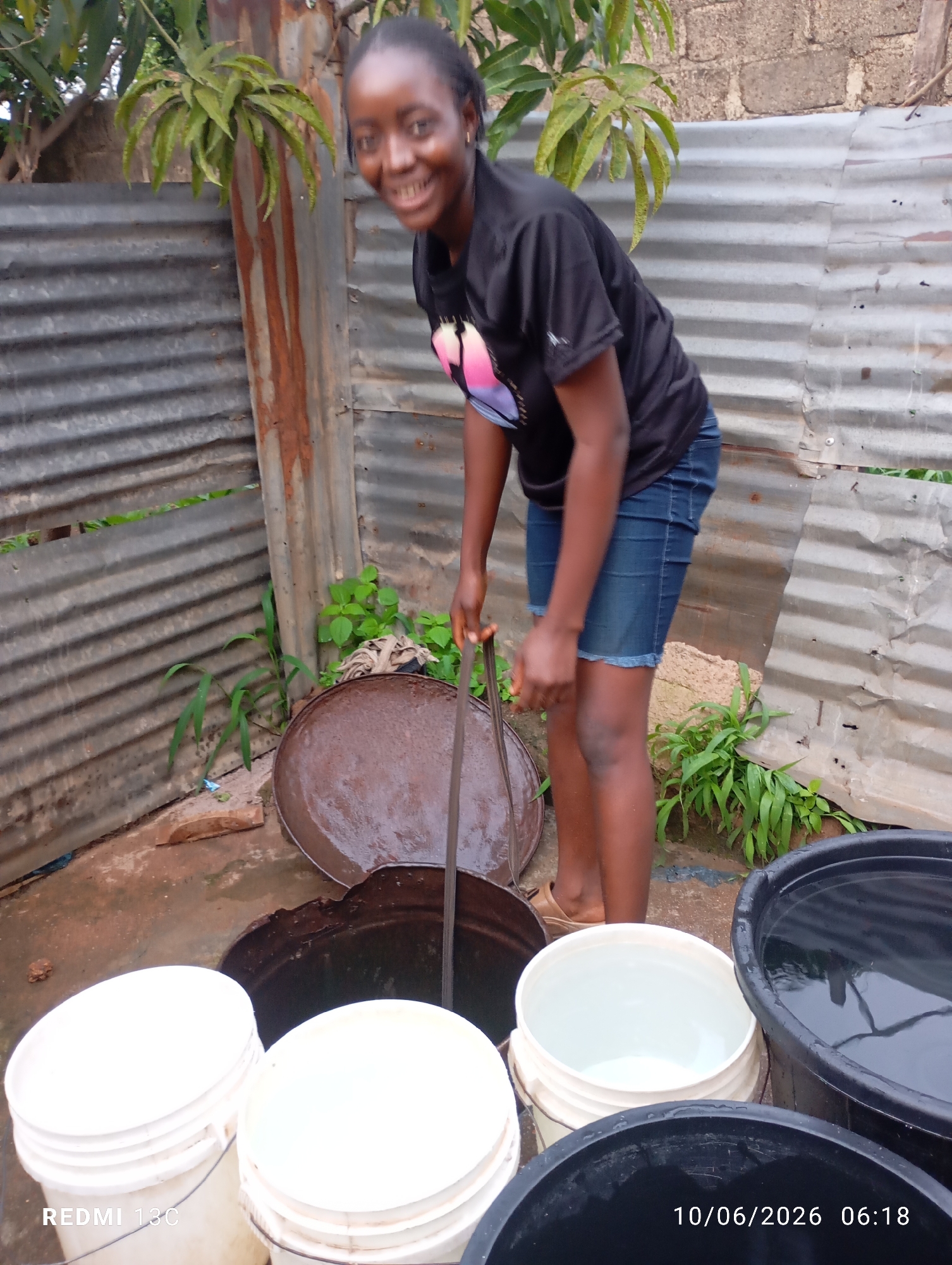 A young Black woman smiles at the camera while holding a pole in a rough outdoor area. She has dark skin and dark hair pulled back. She is wearing a black t-shirt with a colorful design on it, and denim shorts. She is standing in what appears to be a courtyard or backyard, with corrugated metal walls and some vegetation visible. In the foreground, there are several buckets, some filled with water and others empty. A large rusty circular object is also present in the scene, possibly a lid or a pan. The ground is uneven and appears to be dirt or mud, with some puddles of water. The overall impression is of a functional, possibly rural or undeveloped, setting where water collection is a task.