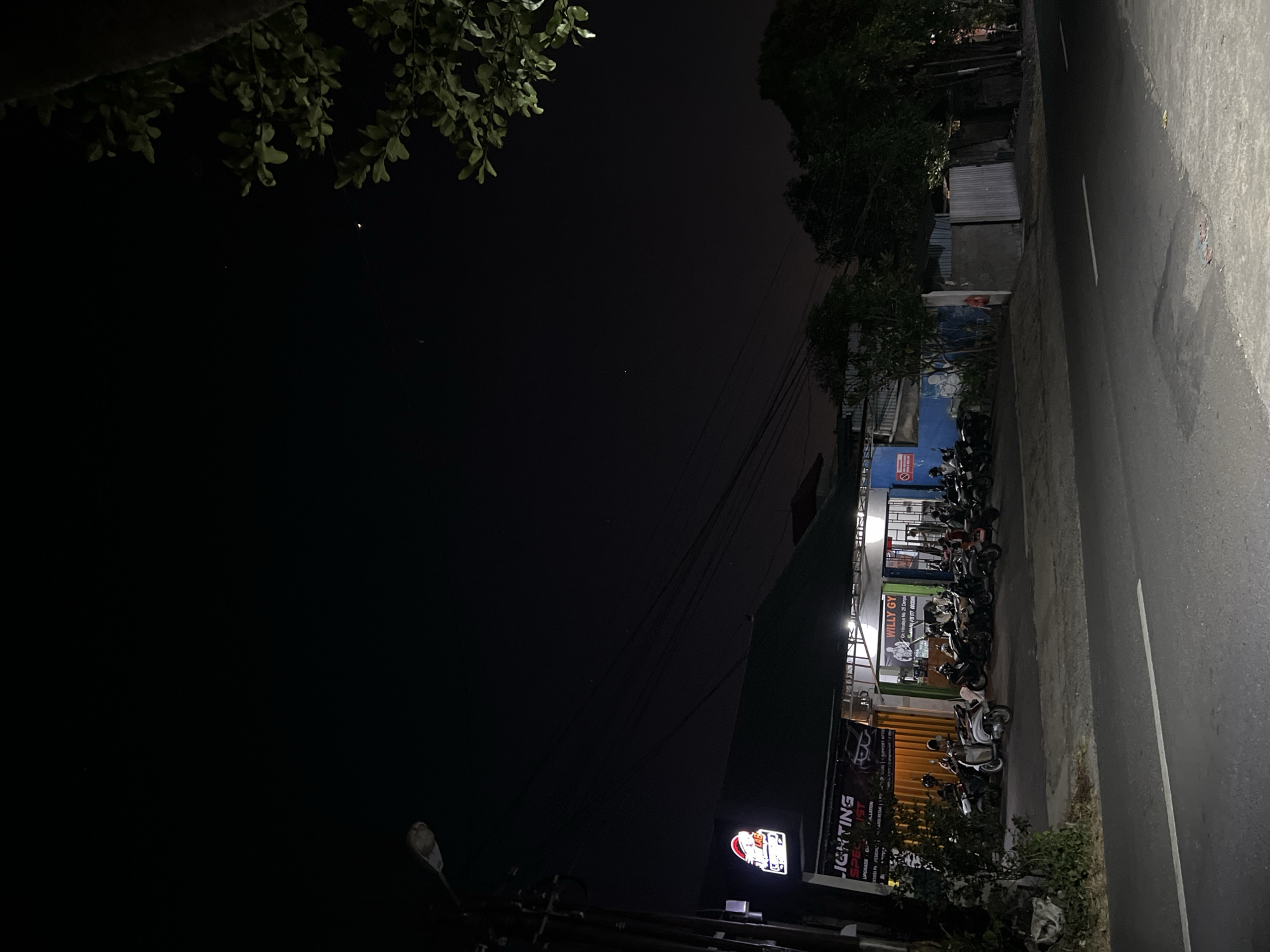 The image captures a nighttime street scene in Denpasar, Indonesia, with a focus on a row of shops and parked motorcycles. A prominent sign on the left reads "LIGHTING SPECIALIST" with a stylized logo. Adjacent to it is another establishment with a sign for "WILLY GYM" featuring an image of a weightlifter. Several motorcycles are parked in front of these businesses. The street in the foreground is asphalt, with white lane markings visible. The sky is dark, with a few faint stars and a sliver of the moon. Trees and a streetlamp are also present in the composition. The overall impression is one of a quiet, commercial area at night.