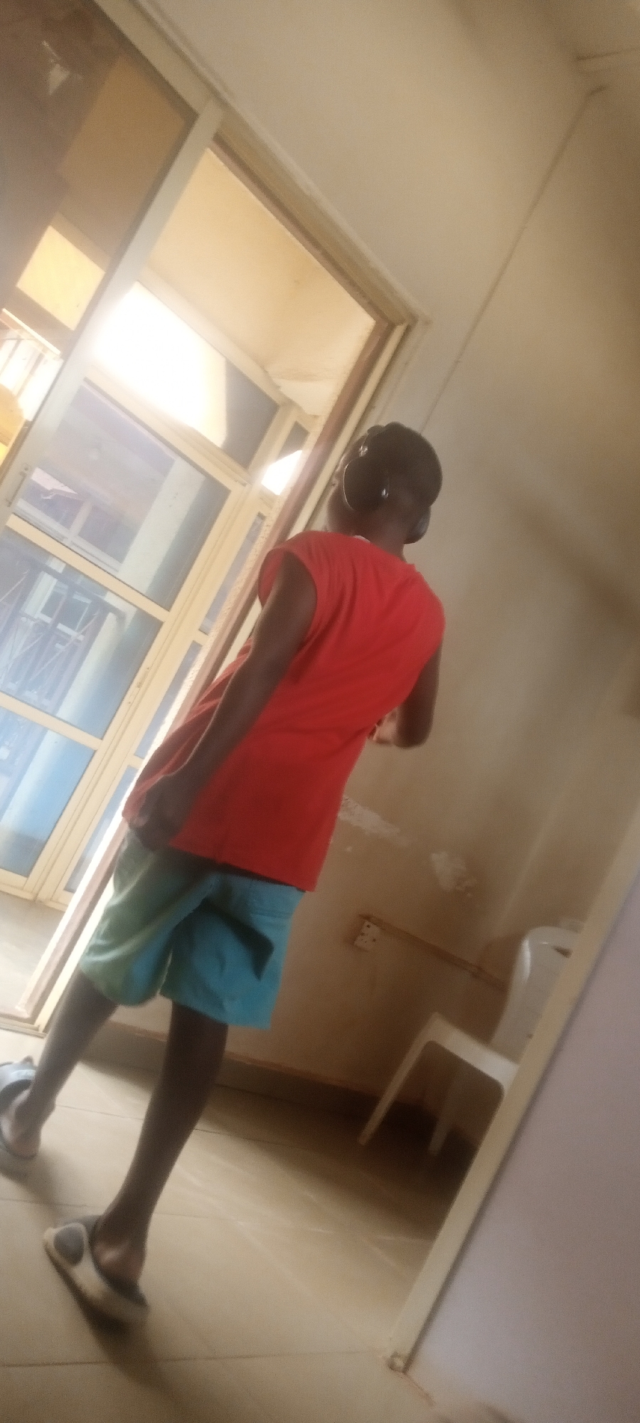 A person wearing an orange sleeveless shirt, blue shorts, and sandals stands in a room near a window. They are also wearing headphones. There's a white chair in the background. The room appears to be in a building in Yelwa, Nigeria.