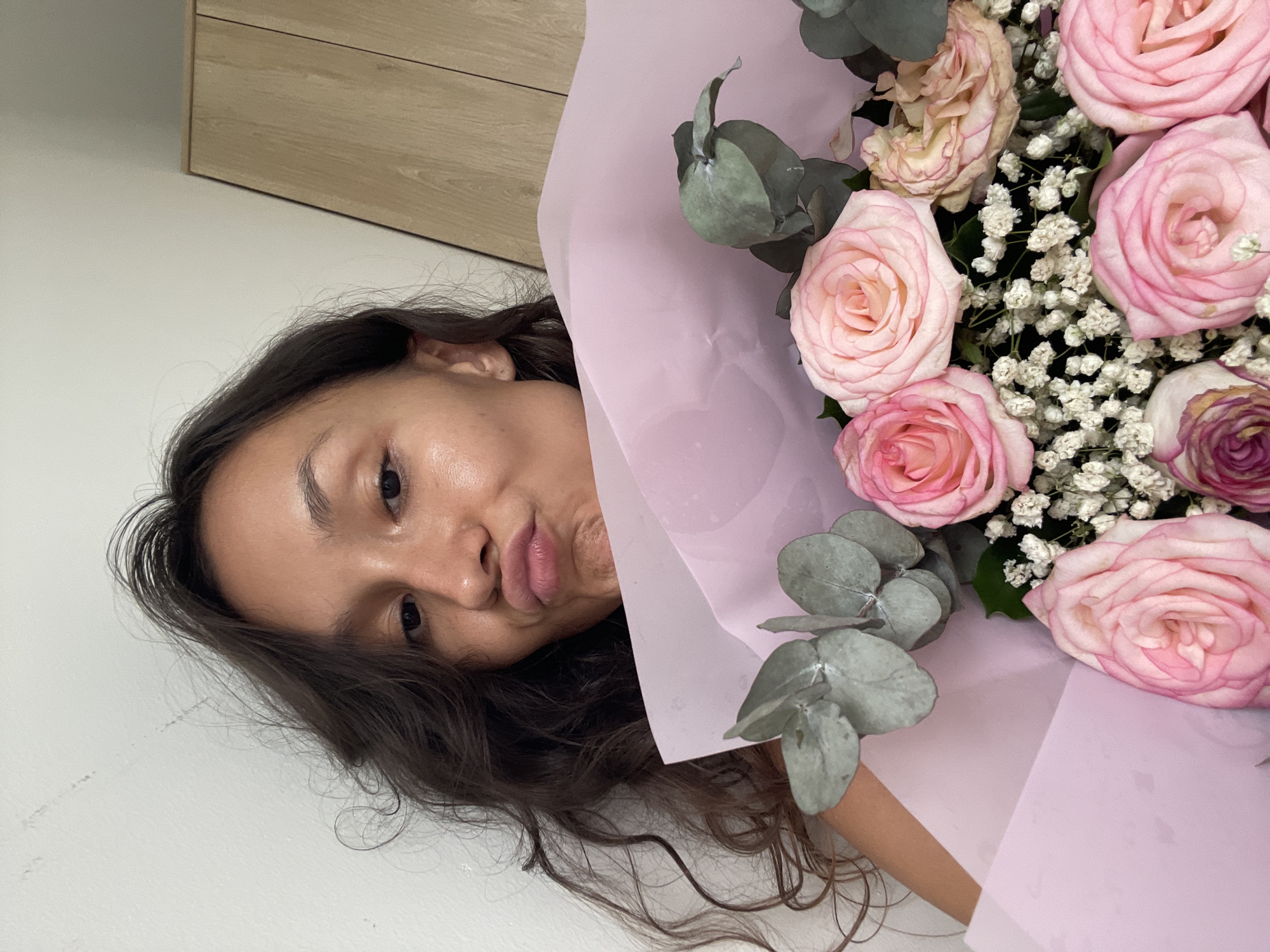 A young woman with long, dark, curly hair, wearing a dark top, is holding a bouquet of pink roses and eucalyptus. She is making a kissing face towards the camera. She is positioned in front of a white wall with some faint marks. To the right of the frame, a wooden closet is visible. The scene appears to be indoors. The woman's location in Phu Thuy Ward, Vietnam, is not determinable from the image.