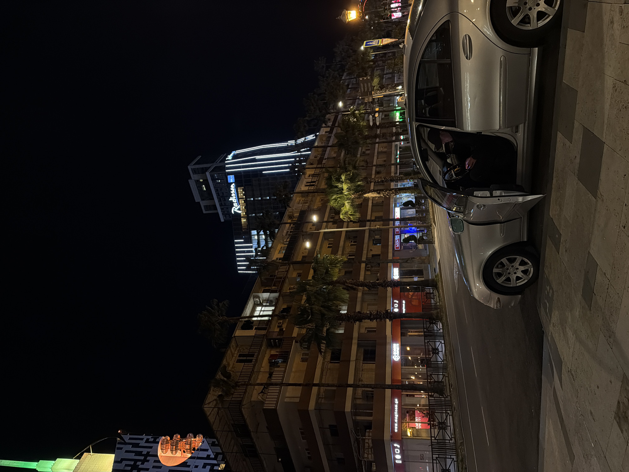 Captured at night in Batumi, Georgia, the image features a silver Mercedes-Benz parked on a sidewalk adjacent to a multi-story building, with its driver-side door ajar, revealing a person inside. The Radisson Blu Hotel, identifiable by its signage and illuminated architecture, stands prominently in the background. The surrounding urban environment includes palm trees lining the street, commercial establishments with lit signs, and streetlights casting a warm glow. The atmosphere suggests a bustling city center at night.
