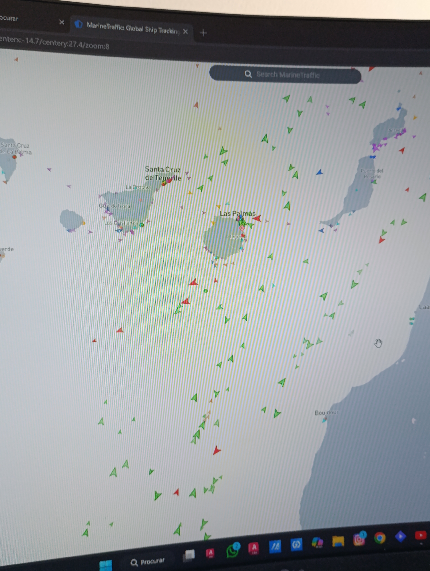 The image shows a computer screen displaying the MarineTraffic website, a global ship tracking service. The map is centered on the Canary Islands, specifically focusing on Tenerife and Gran Canaria. The cities of Santa Cruz de Tenerife, La Orotava, Gran Canaria, Los Cristianos, Las Palmas, and Ingenio are labeled. The map displays numerous arrow symbols representing ships; these symbols are colored red, green, blue, and purple, indicating different types of vessels or their status. To the east is the city of Puerto del Rosario. To the south is the city of Boujdour. The top of the screen shows the browser tab for the MarineTraffic website, along with the URL and zoom level. The bottom of the screen reveals the taskbar of a computer, displaying various application icons, including a search bar labeled "Procurar".