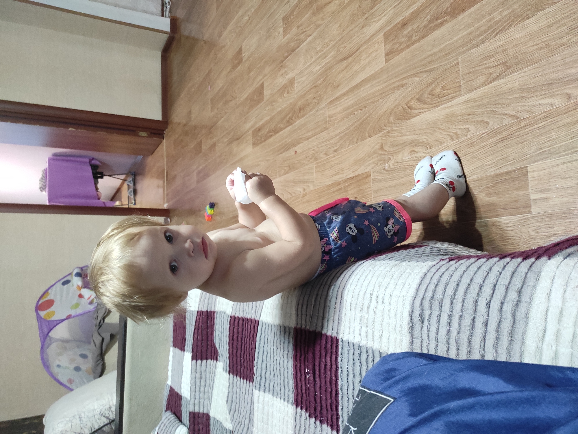 A fair-skinned toddler with blonde hair is sitting on the edge of a bed in a room. The toddler is facing left and looking towards the camera with a sad expression, holding a white tissue in their hands. The toddler is wearing patterned shorts and white socks with cherry designs. The bed has a striped comforter with shades of grey, white, and maroon. To the left of the bed, a blue garment is partially visible. The floor is made of light brown laminate. In the background, there is a doorway leading to another room where a table is covered with a purple cloth. Further in the background, a purple pop-up play tent with colorful polka dots is visible. The scene appears to be indoors, possibly in a residential setting. The location is Shakhtinsk, Kazakhstan.
