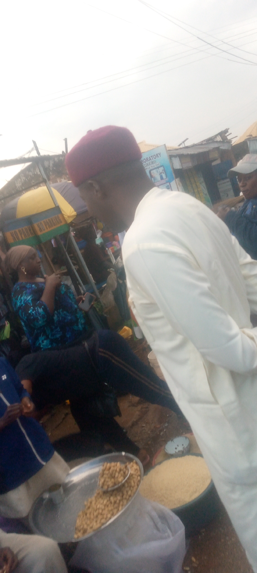 In an outdoor market setting, a person wearing a maroon kufi and a light-colored robe is seen from the side, facing away from the camera. To the left, a woman in a dark blue and black patterned top and dark pants sits, looking to her left. She is wearing a headscarf and earrings. A smartphone is visible in her hand. In the foreground, a silver platter contains a pile of small, light brown nuts, with a spoon resting on top of them. Beside the platter, a large black basin is filled with a mound of pale yellow grains, possibly rice. Another person in a dark jacket and a cap is visible in the background to the right. Behind the people and market stalls, power lines are visible against an overcast sky. Signs are present on some of the market stalls, one of which reads "BORATORY". The ground appears to be dirt or mud.