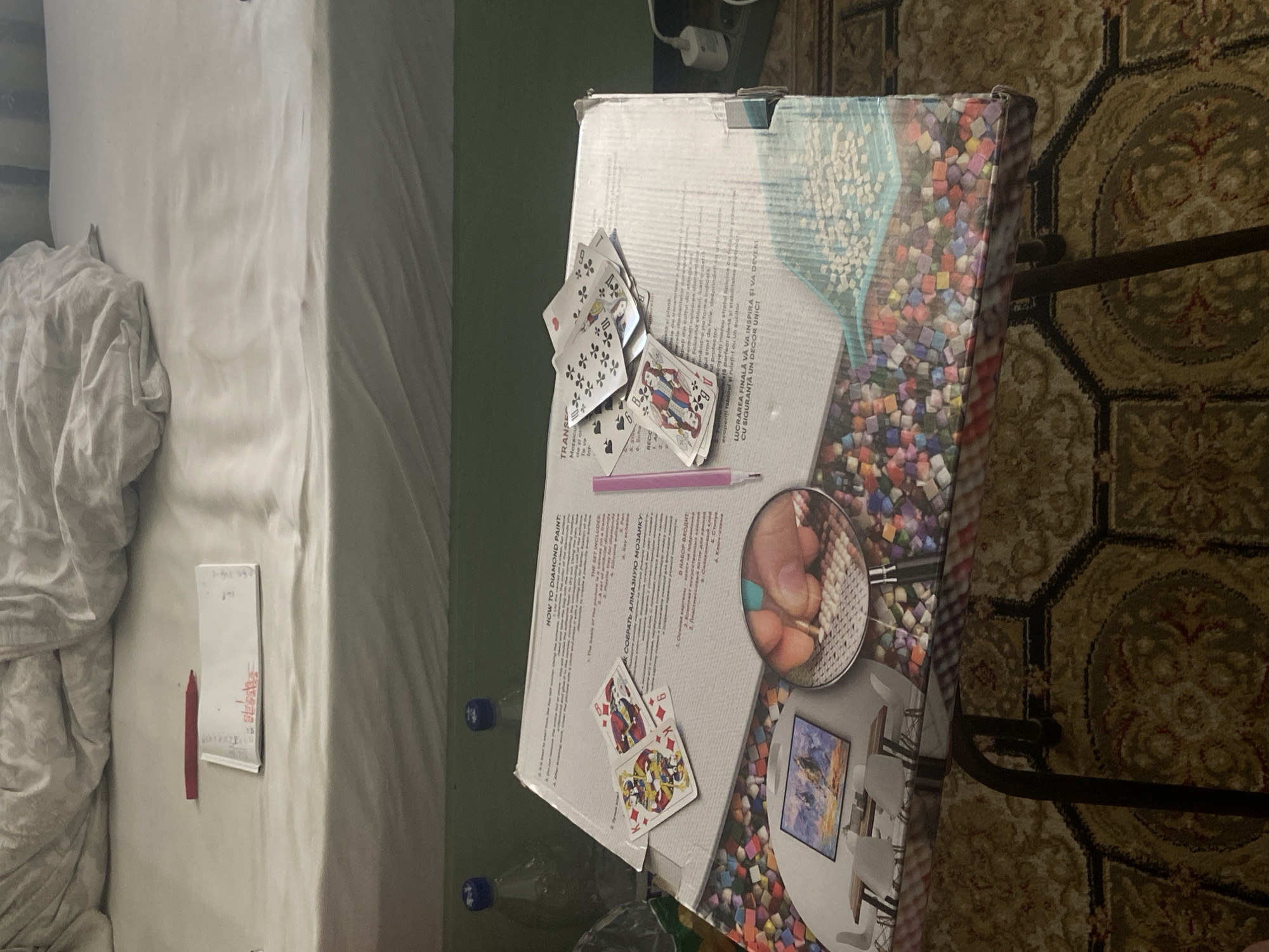 The image shows a flat lay of objects on top of a cardboard box which is on top of a dark metal folding table. The cardboard box is partially covered with playing cards, and has text and images printed on it. There are also two bottles of water in the background, and the scene is set on a bed with white sheets. The overall impression is that of an indoor scene, likely a bedroom or living space, with a focus on the game or activity represented by the playing cards and the box.