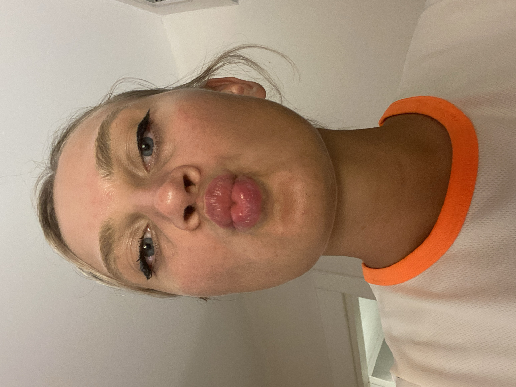 The image shows a close-up, front-facing selfie of a young woman with blonde hair and blue eyes, puckering her lips. She is wearing a white t-shirt with an orange trim around the neckline. The lighting suggests an indoor environment. There is no visible indication of the city of Aksu, Turkey, or any urban elements.