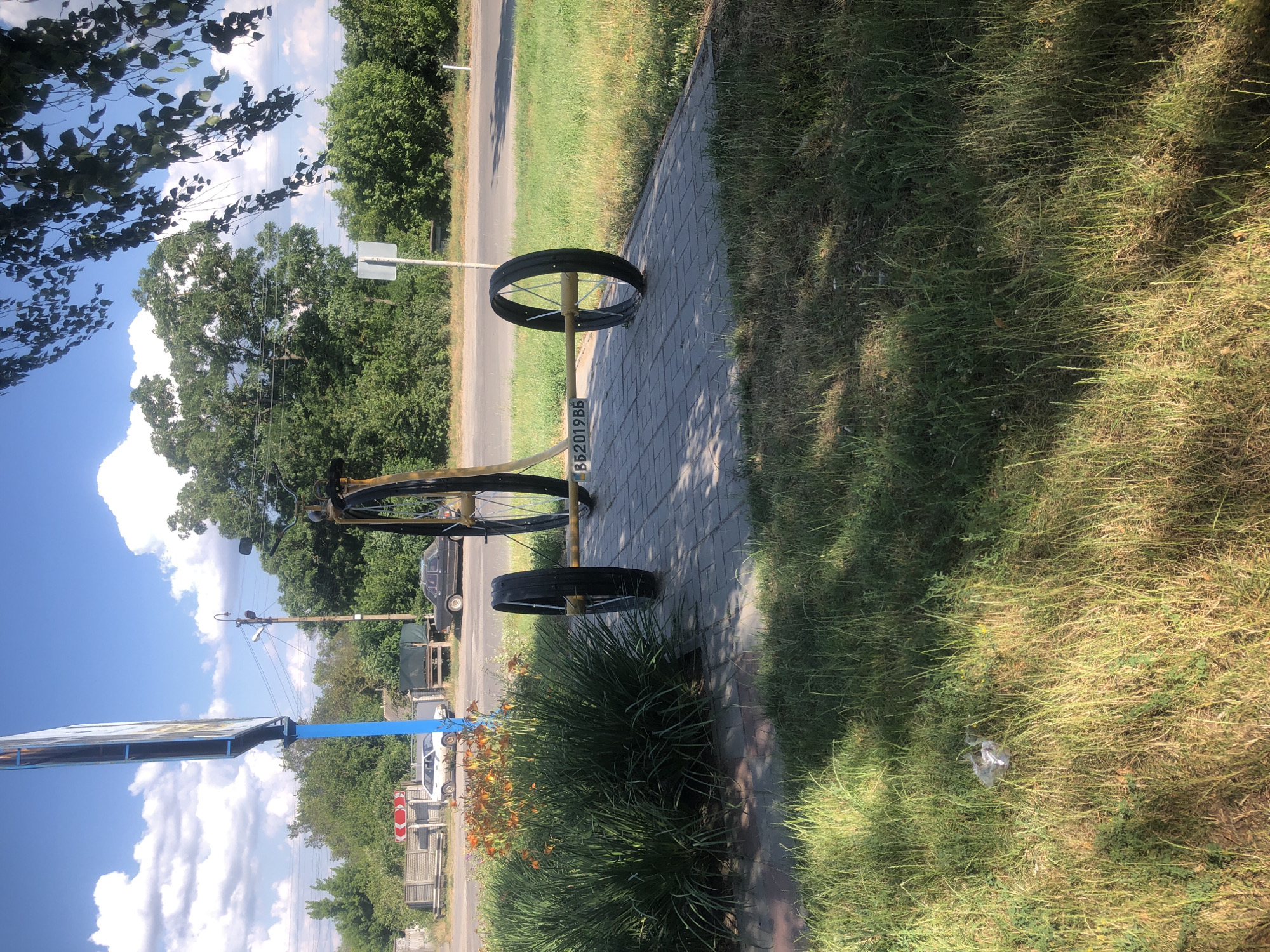 The image captures a scene outdoors, likely during daytime, given the bright sunlight casting shadows and the clear blue sky with scattered clouds. The primary object of focus is a yellow metal frame with two large black bicycle wheels, suggesting a wheeled apparatus or a part of one. A license plate is attached to the yellow frame, displaying the text "BB2019B5". The apparatus rests on a paved walkway made of grey rectangular bricks, which runs alongside a grassy area.
In the background, a road can be seen, bordered by more grass and trees. Several vehicles are present: a dark-colored car parked on the side of the road, and a white car further down. There are also utility poles and some fencing visible, hinting at a semi-rural or suburban setting. A large blue signpost with a white rectangular panel is also in the frame, but the content on the panel is not clearly discernible. The presence of lush green vegetation, including trees and bushes, suggests it's a warm season. The overall impression is a still, outdoor scene, possibly a roadside or public area, with no people immediately visible.