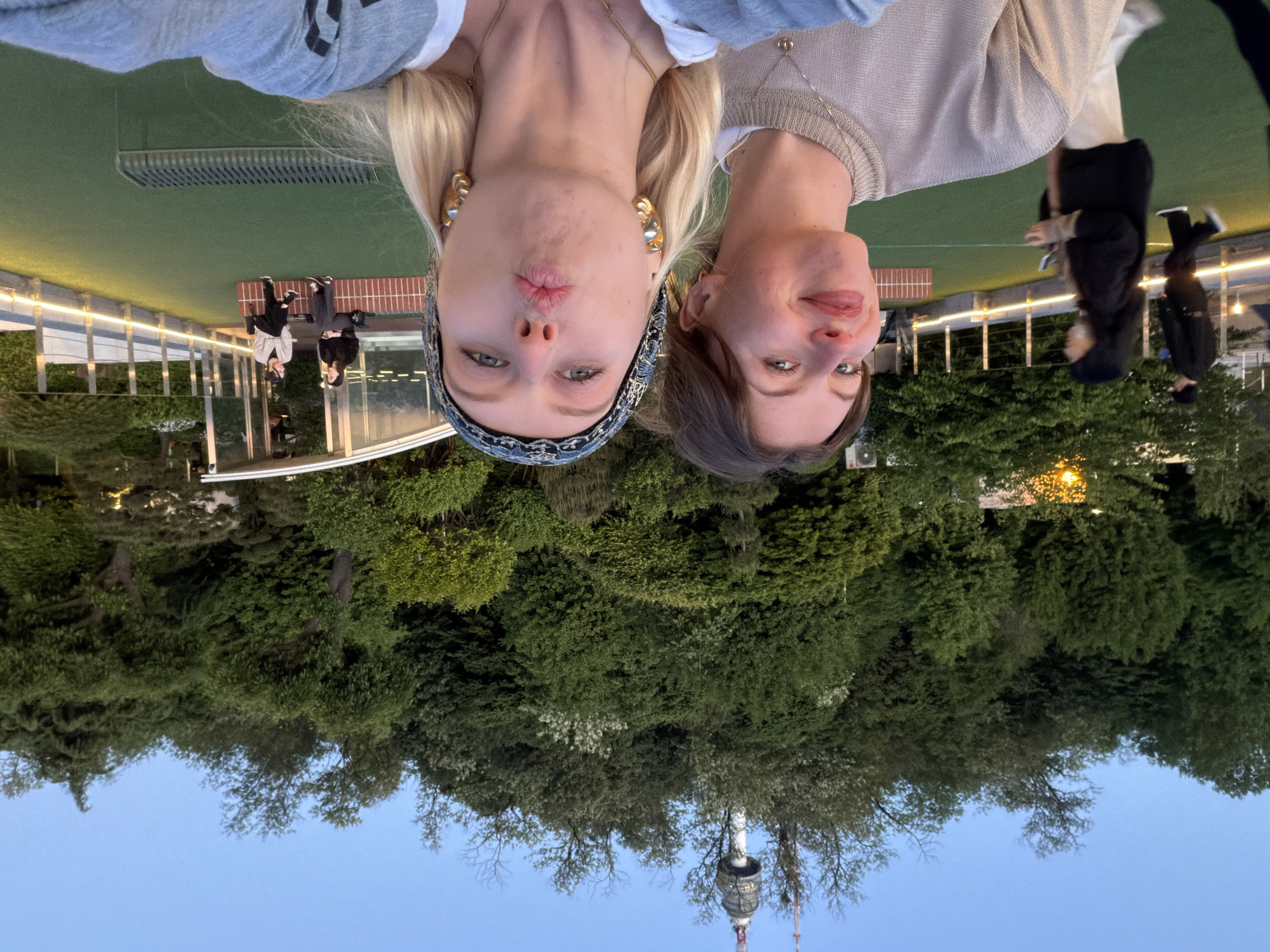 Two women are posing in a selfie with the Seoul Tower visible in the background. The woman on the left has blonde hair and is wearing a patterned headband and large hoop earrings. The woman on the right has brown hair and is also wearing a necklace. They are standing on a platform surrounded by trees. In the background, there are benches and other people, likely visitors to the Seoul Tower area. The sky is a clear light blue.
