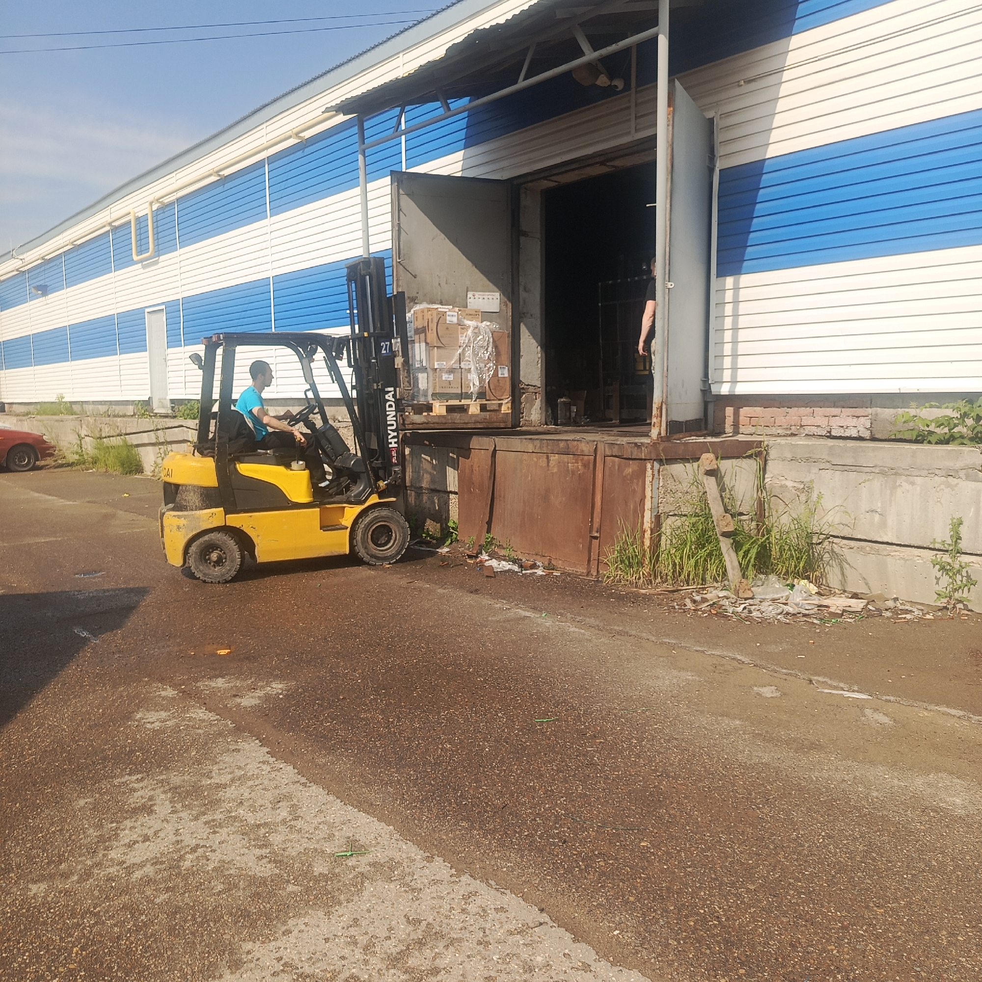 The image depicts a yellow Hyundai forklift operating outside a warehouse with blue and white striped siding. A man in a bright blue t-shirt is driving the forklift, which is positioned near a loading dock. Pallets of boxes are visible inside the open warehouse door, some of which appear to be wrapped in plastic. The ground is asphalt with some debris. A red car is parked to the left of the scene. The sky is clear and blue. The overall impression is of an industrial or commercial area. This location is in the city of Chaikovsky, Russia, a city known for its industrial enterprises, particularly in the chemical and petrochemical sectors, as well as energy production.