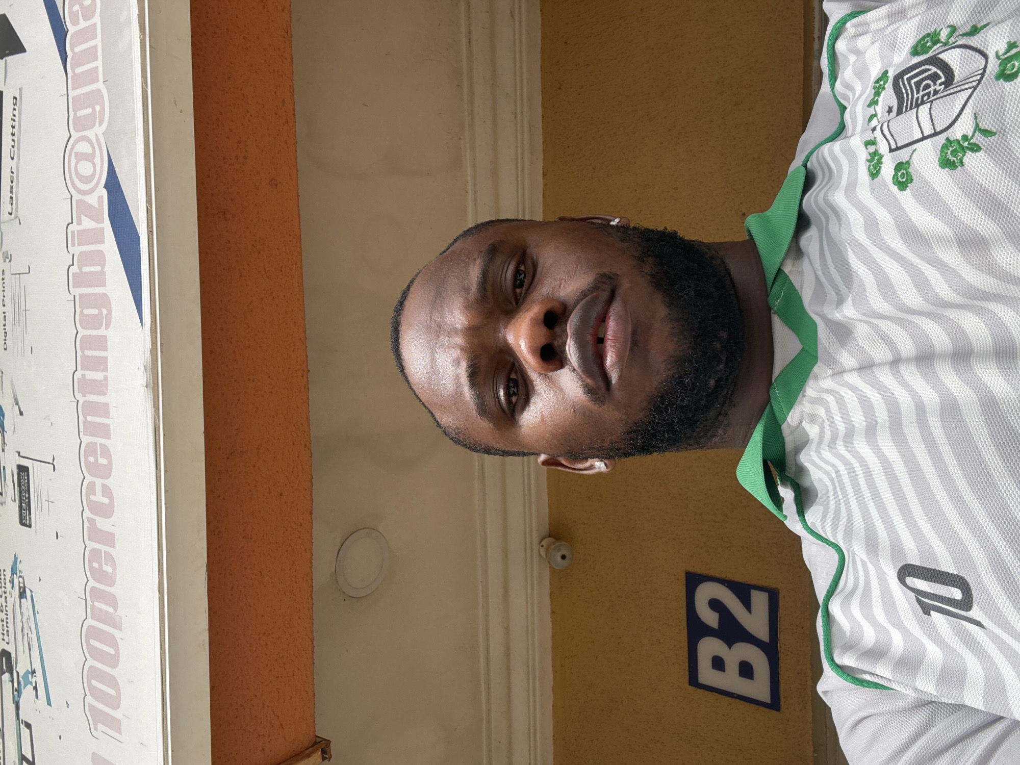 A close-up, rotated, outdoor photo shows a dark-skinned man with a beard and short hair wearing a white and grey striped soccer jersey with green trim and the number "10" on it. Behind him is a mustard-yellow wall with a "B2" sign and a security camera mounted on the wall. To the left is an advertising sign that says "100percentngbiz@gmail.com" and "Laser Cutting". The location is Lugbe, Nigeria.