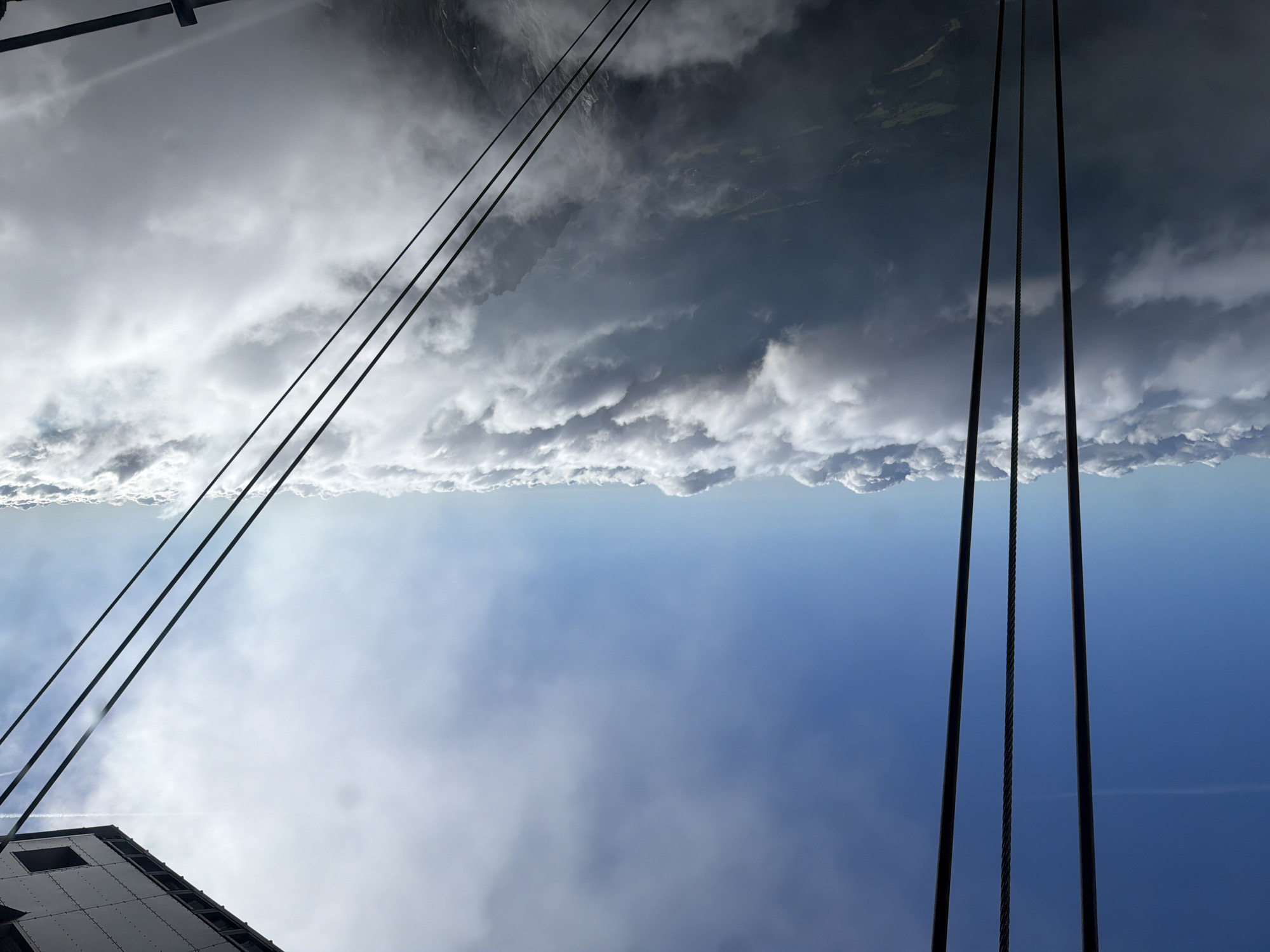 The image shows a view from a cable car traveling above the clouds, likely en route to the Zugspitze, Germany's highest peak, near Garmisch-Partenkirchen. Several thick, dark cables dominate the foreground, running vertically and diagonally across the frame, suggesting the perspective of someone inside or near the cable car. The cables are suspended against a bright blue sky, which is partially obscured by a thick blanket of white, fluffy clouds that stretch across the horizon. Through gaps in the clouds, glimpses of a dark, mountainous landscape with hints of green fields and possibly a village are visible, indicating the descent or ascent over a valley. The sun is shining, casting a bright light that creates some glare and illuminates the clouds from above. The top right corner of the image reveals a section of the cable car's structure, a metallic ceiling with visible rivets and a square panel. The overall scene conveys a sense of height, vastness, and an awe-inspiring natural landscape viewed from an elevated position.