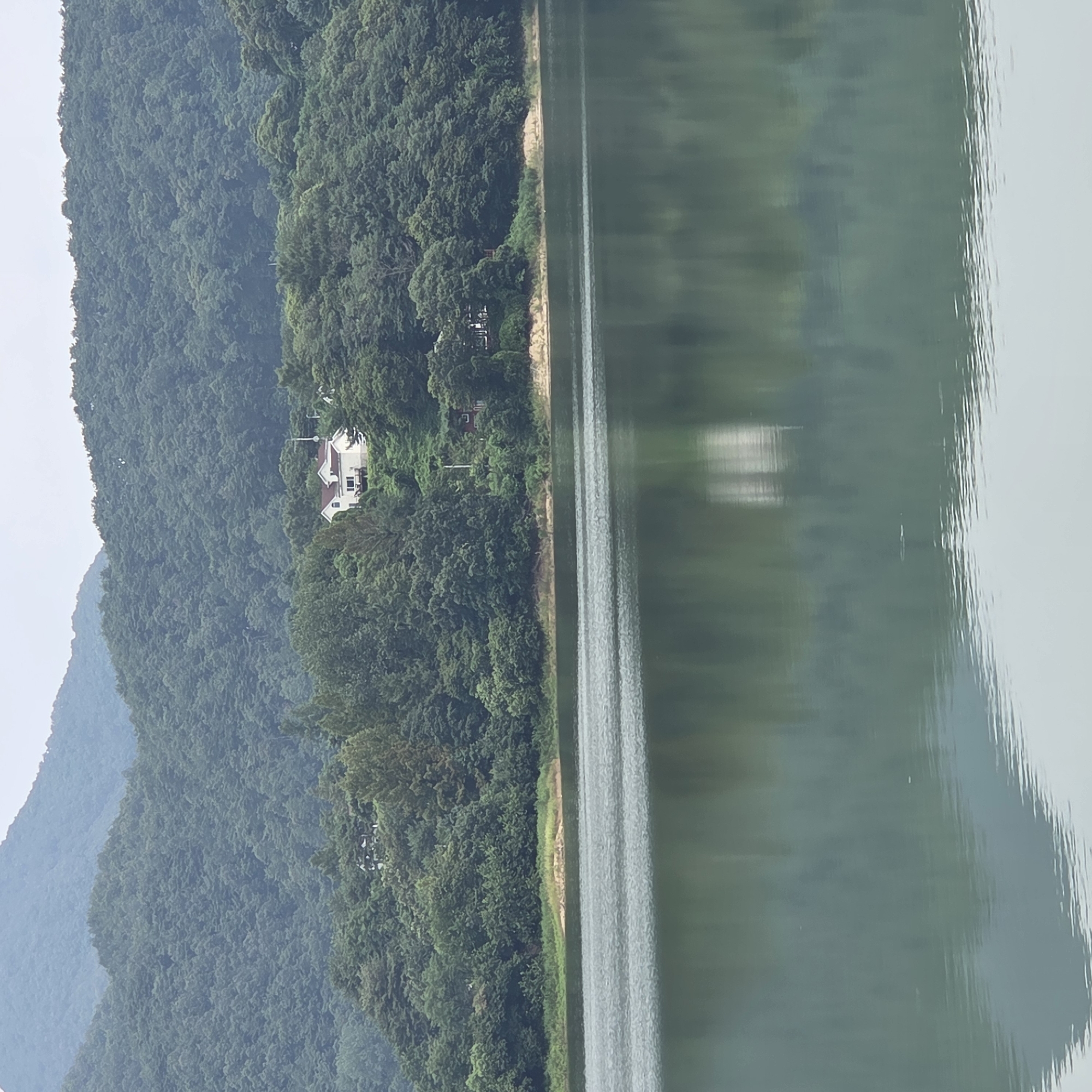 A wide shot captures a serene lake in Gapcheon-myeon, South Korea, with a backdrop of lush green mountains and a dense forest bordering the water. The lake's surface is calm, reflecting the verdant trees and the overcast sky. On a slope overlooking the lake, nestled among the trees, is a modern house with white walls, a dark roof, and a visible balcony. The scene appears tranquil and uninhabited, conveying a sense of natural beauty and rural seclusion.